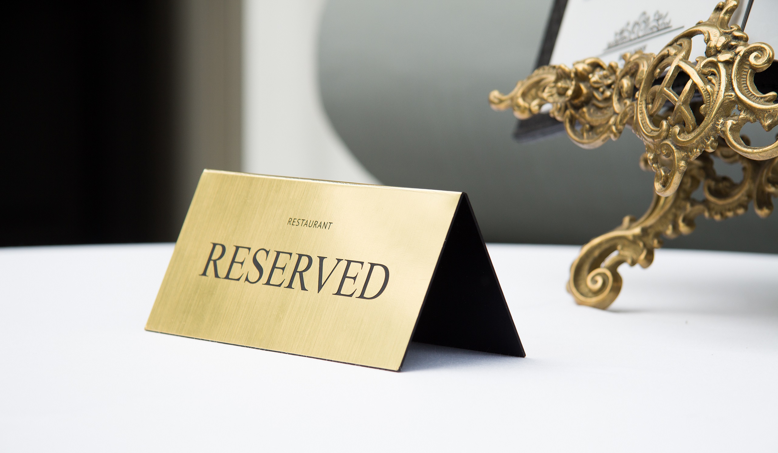 A gold-plated, engraved reserved sign on a restaurant table covered with a white tablecloth also seeing the bottom of an easel picture stand.