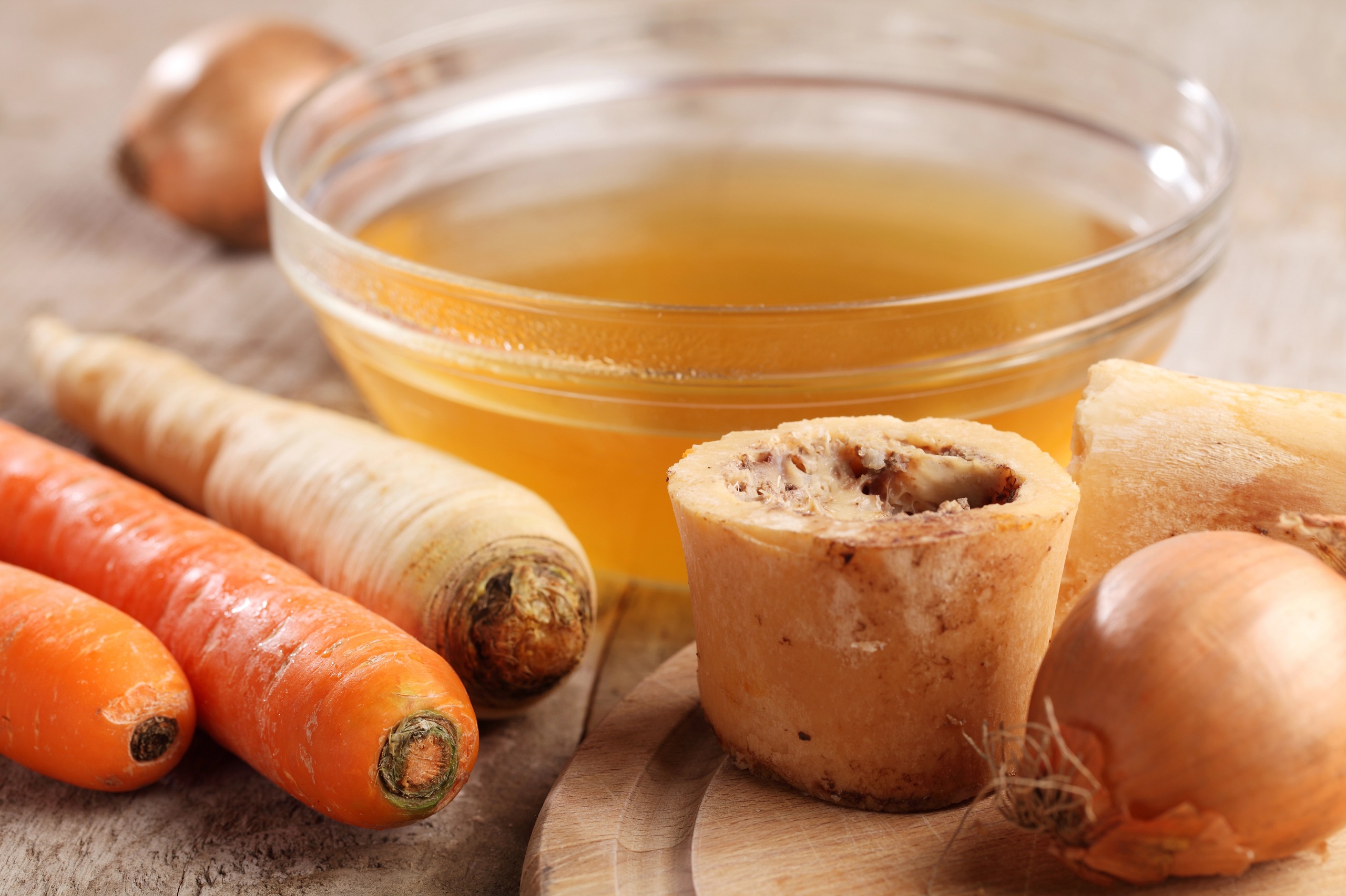 A small clear glass bowl filled with beef broth centered around a couple pieces of boiled bones with visible marrow next to orange and white carrots and yellow onions on a wooden surface.