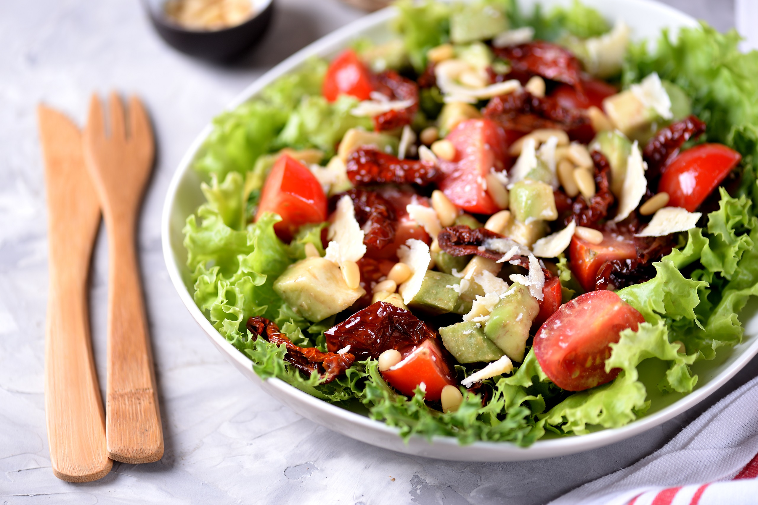 Berries, nuts, tomatoes, avocado, and mixed greens salad served in a white salad bowl on a clean, white tablecloth with a traditional wooden fork and knife at its side.