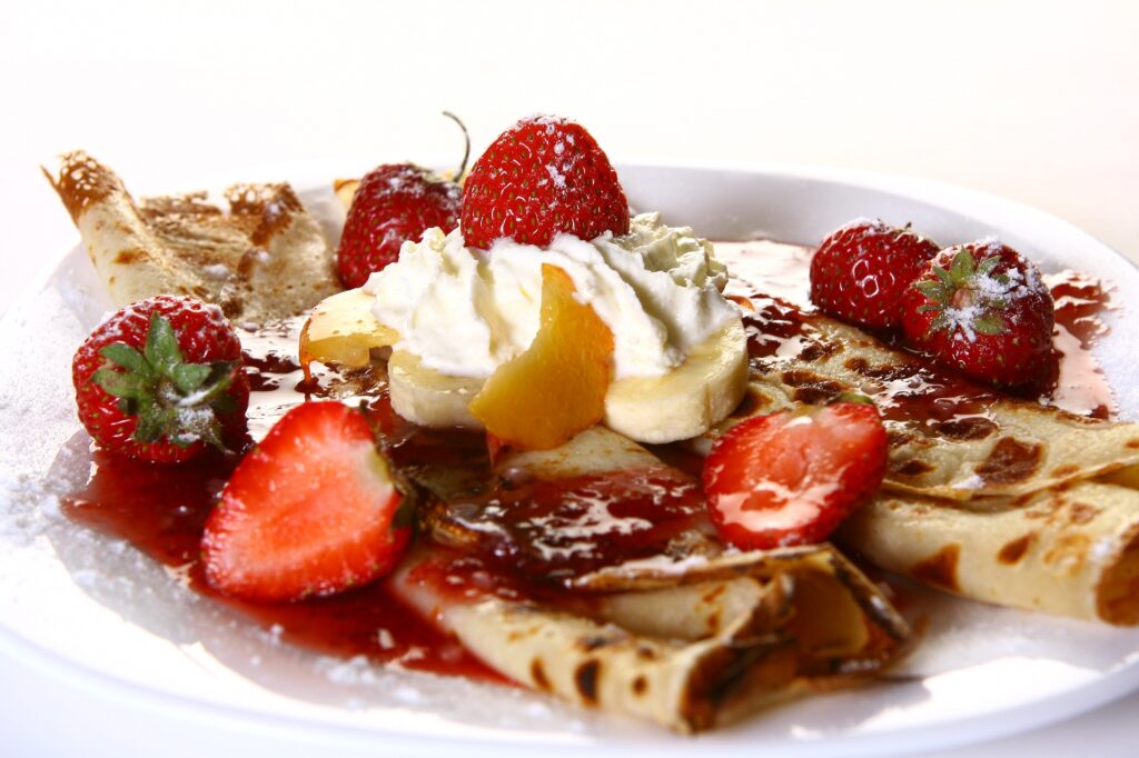 Blintzes with homemade chunky fruit sauce and homemade whipped cream garnished with fresh whole and slice strawberries, sliced bananas, and sliced peached on a white plate sprinkled with powdered sugar
