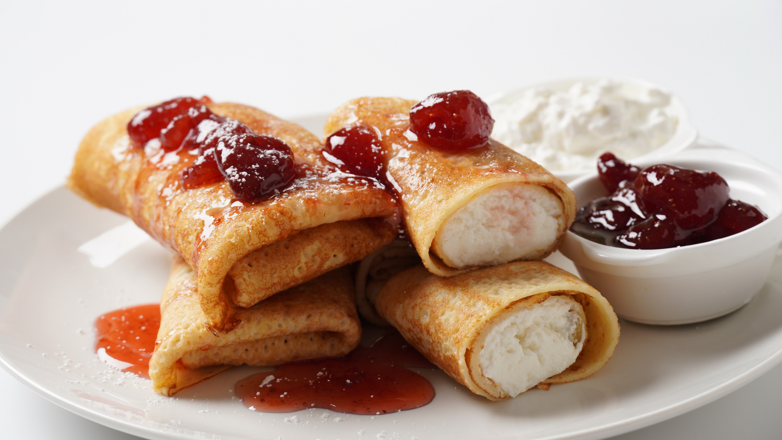 Sweet cheese blintzes stacked on a white spread with the cheese filling showing and natural fruit jam spread across the top row of blintzes on a white background.