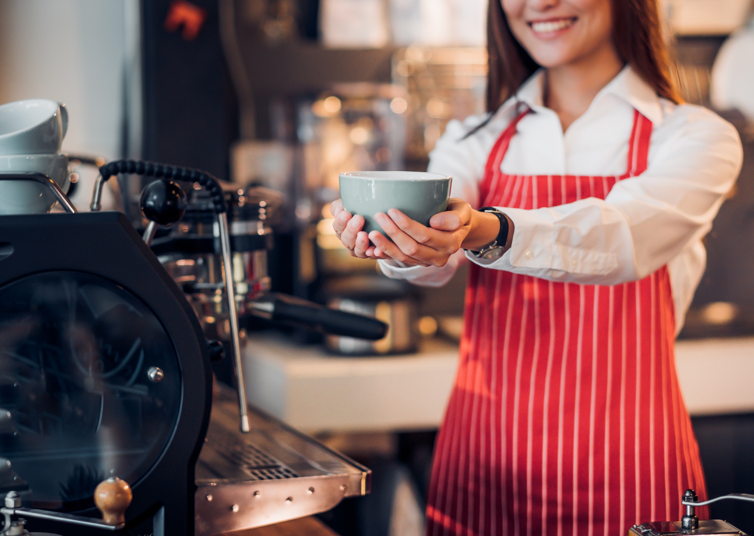 Close up a woman wearing a red and white striped apron cupping a coffee mug with both hands extended as if offering it to someone with a coffee shop in the background conceptualizing a free hot coffee.
