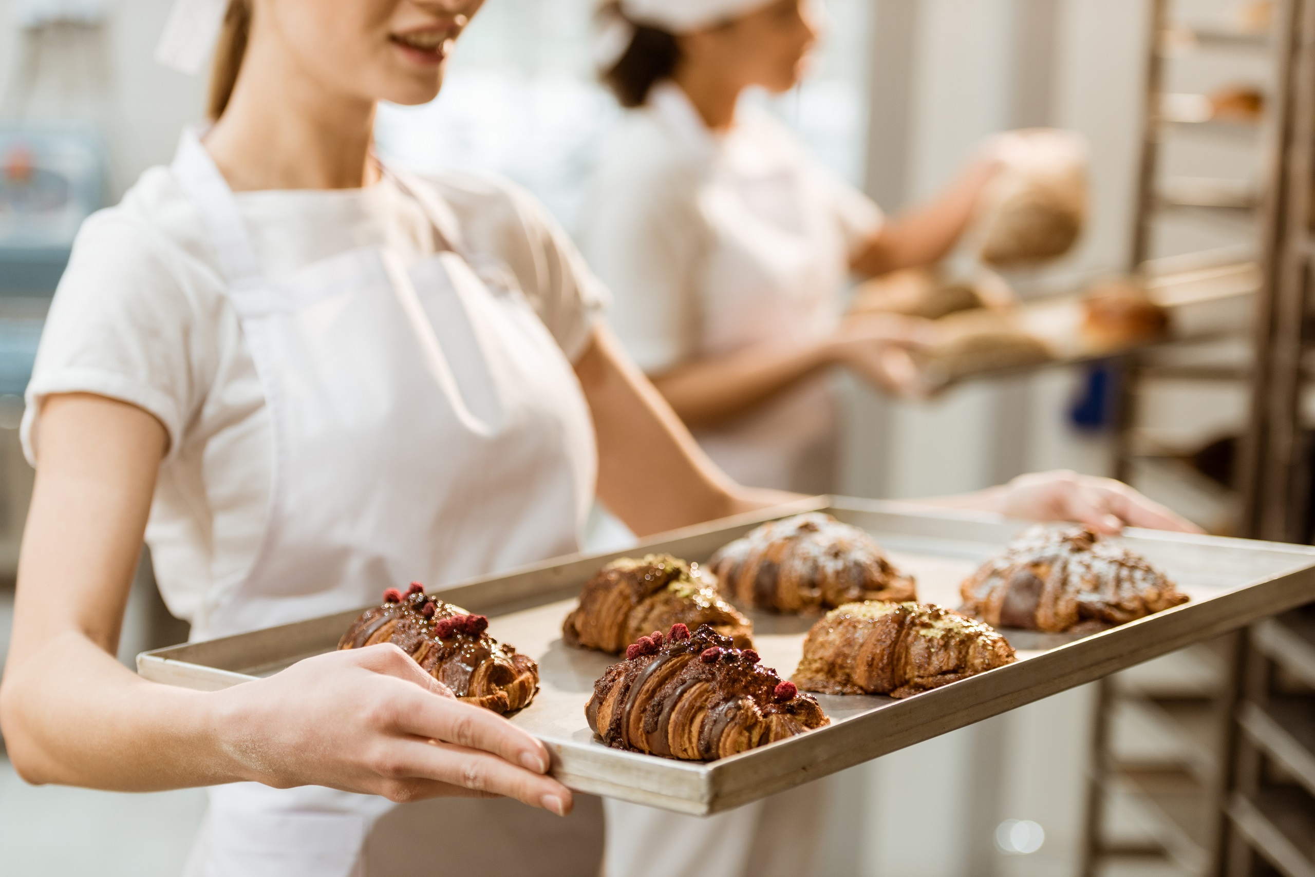 Cropped shot of young female baker holding a tray of fresh gourmet croissants with another female baker in the background pulling baked goods off of a rack.