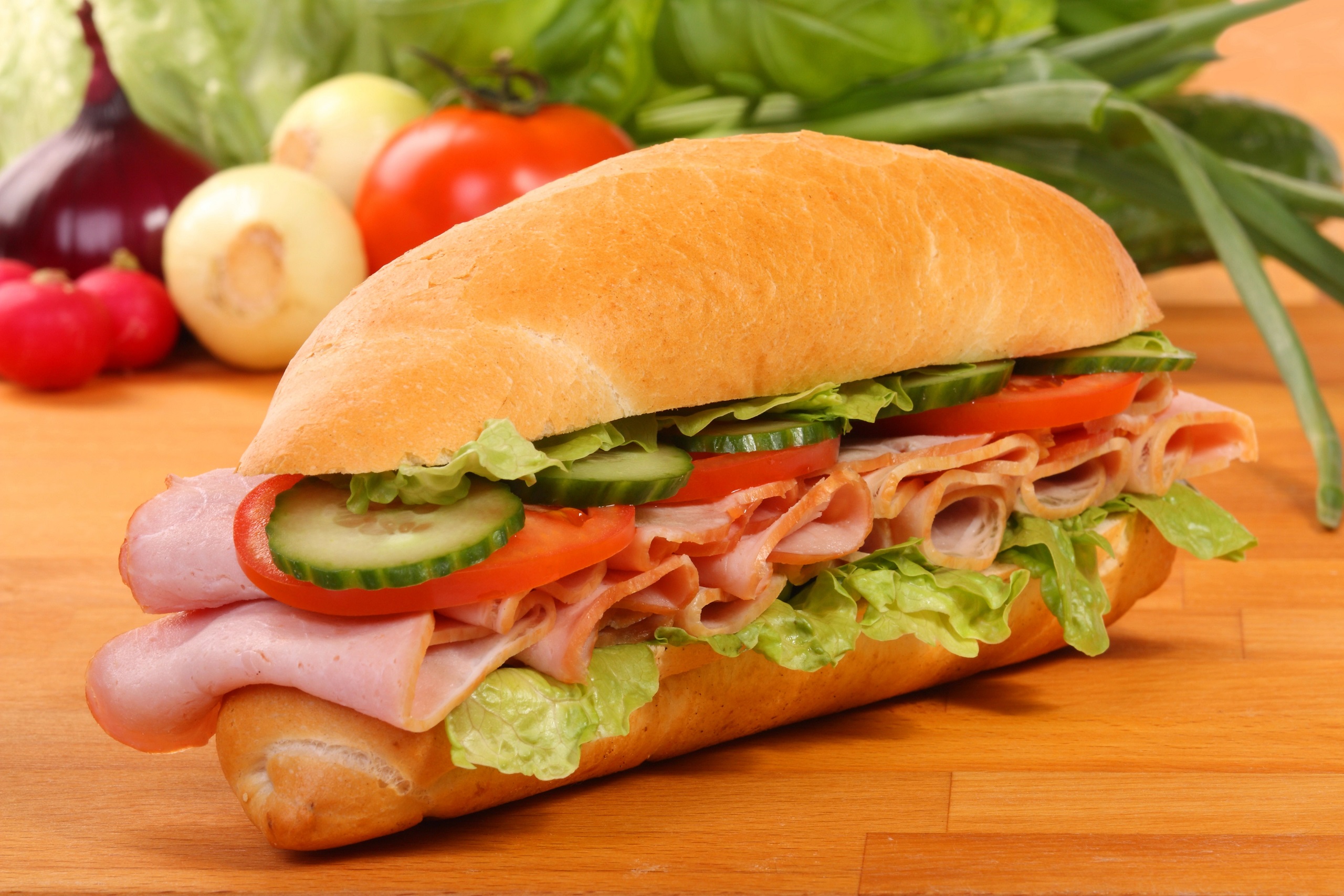 Delicious fresh gourmet sandwich packed with off the bone ham, swiss cheese, lettuce, tomatoes, cucumbers, and other vegetables on a wooden tray.