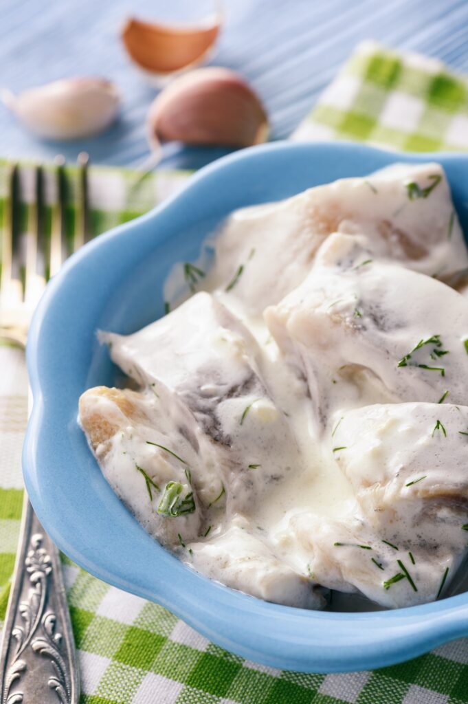 Pieces of marinated herrings made in creamy dill sauce served in a blue bowl on a green and white checkered napkin on blue surface with a engraved silver fork and garlic bulbs next to it.