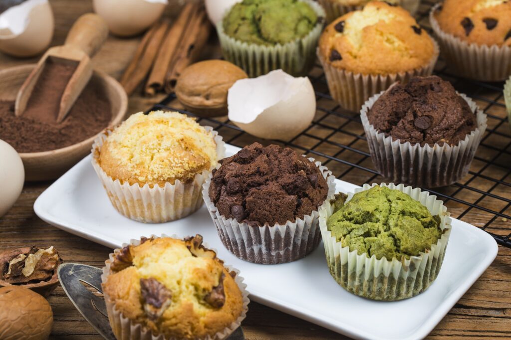 A variety of fresh muffins such as chocolate fudge, blueberry, pistachio, banana, chocolate chip loseely arranged on a brown and white surface along with cracked egg shells, walnuts, and cinnamon sticks.