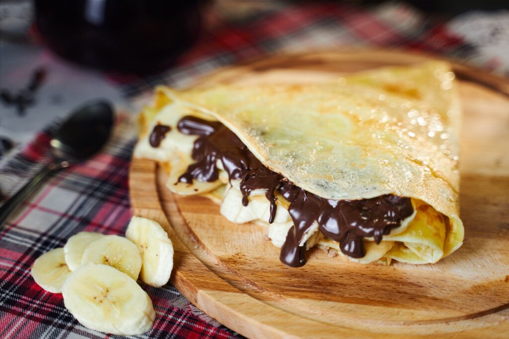 Nutella and banana bltinz folded into a triangle on a wooden plate with sliced bananas to the side of the plate on a luxuriously woven plaid tablecloth.