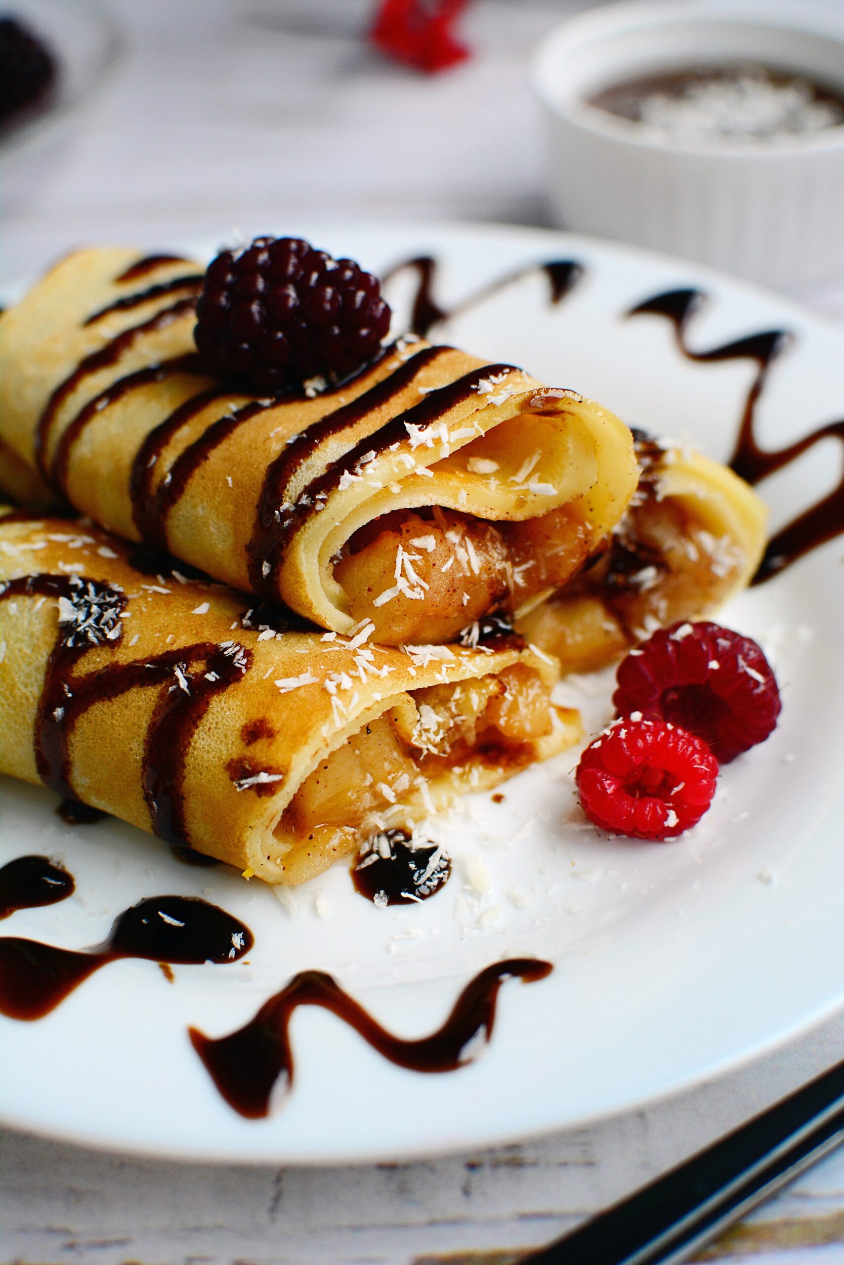 Peanut butter and fruit jam blintzes with rasberry and blackberry garnish sprinkled with cococunt or powdered sugar flakes on a white plate further garnished with swirls of chocolate syrup.
