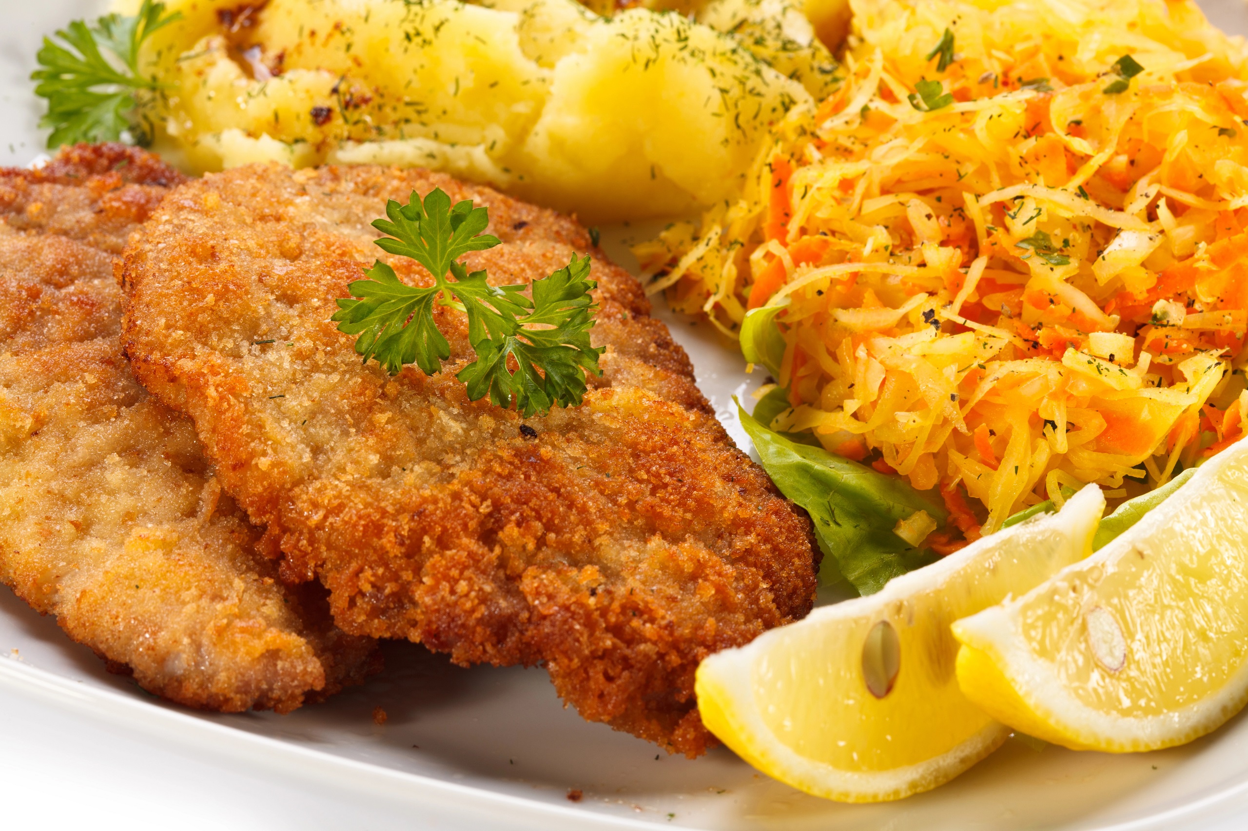 Breaded pork cutlet with mashed potatoes and sauerkraut salad on a white plate garnished with parsley and lemon wedges.