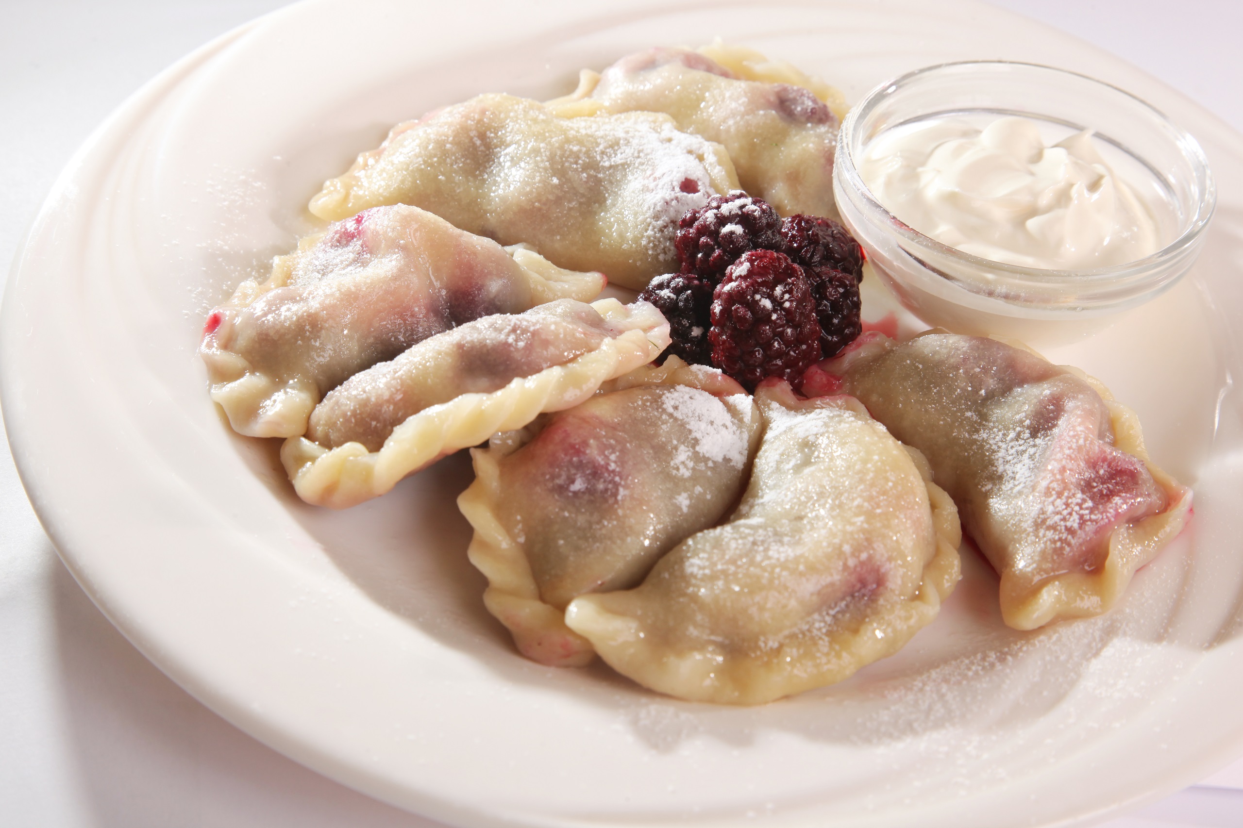 Several pierogies with rasberry filling, topped with rasberries and sprinkled with sugar, neatly arranged on a plate served with a small side of sour cream.