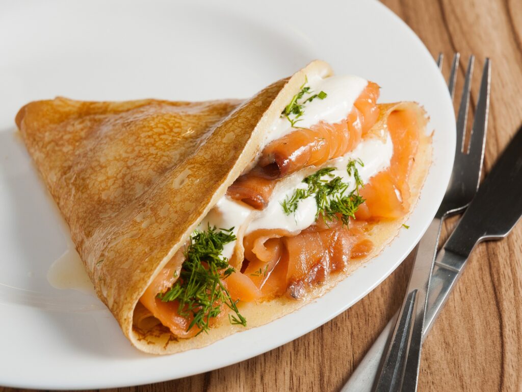 A folded blintz filled with thinly sliced smoked salmon and cream cheese with dill garnish on a plate next to a fork and knife on a wood grain surface.