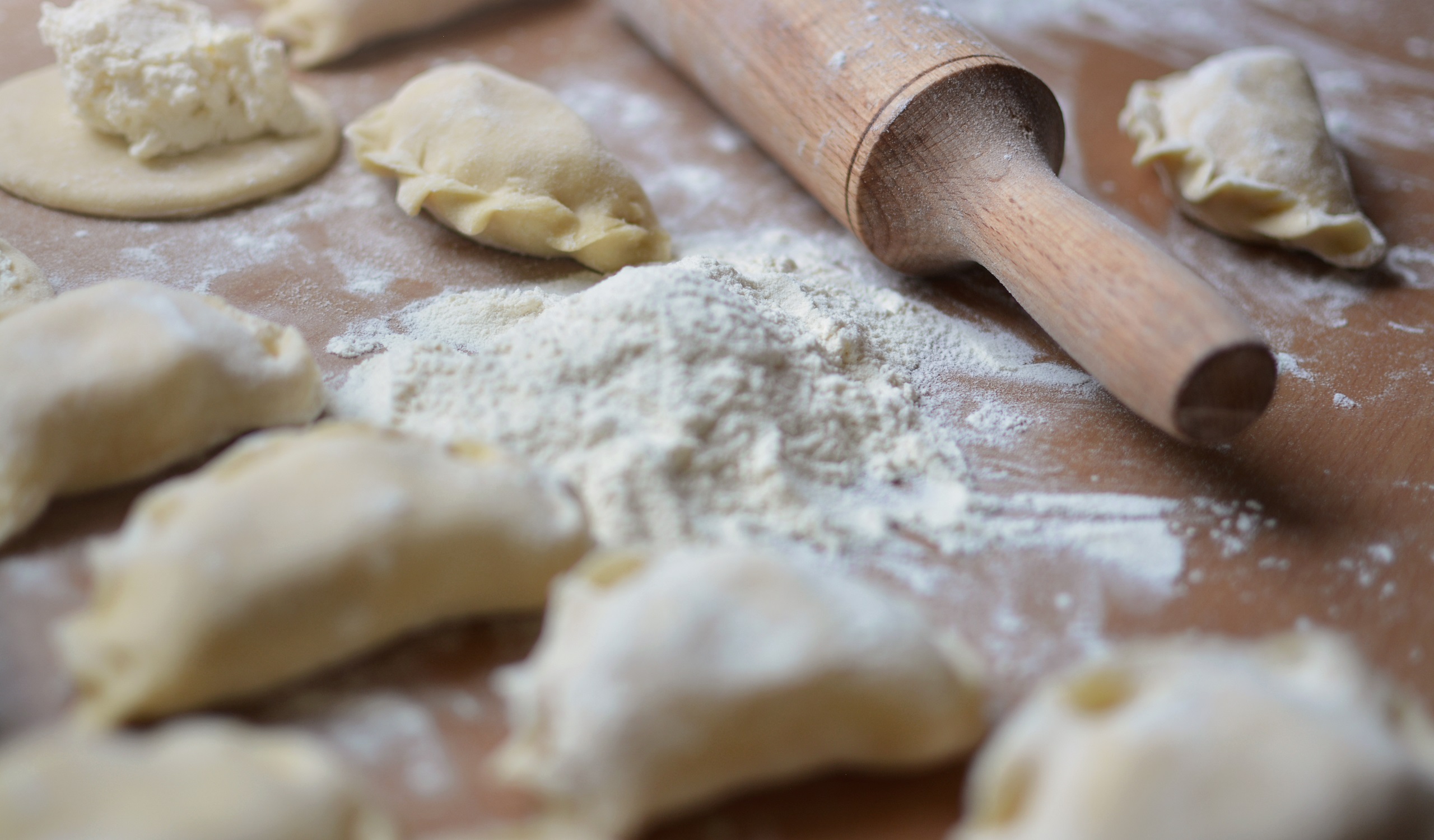 Different steps in the process of making pierogies from a small pile of flour, a small flat pierogi dough having a pinch of filling at its center, several finished raw pierogies, and a wooden rolling pin.