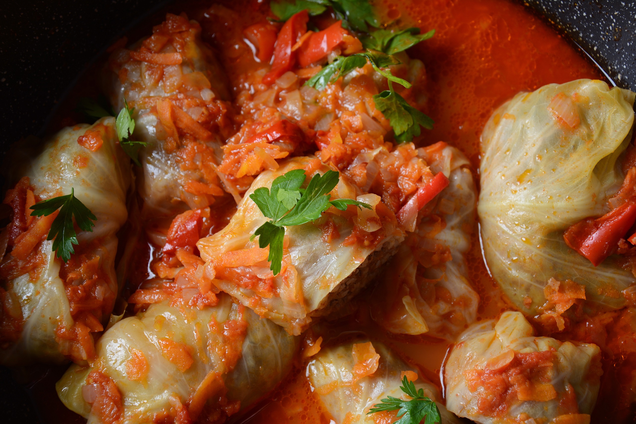 Close up of stuffed cabbage rolls smothered in chunky light tomato sauce with carrot shreds garnished with parsley still in their juices from when cooked in an iron skillet.
