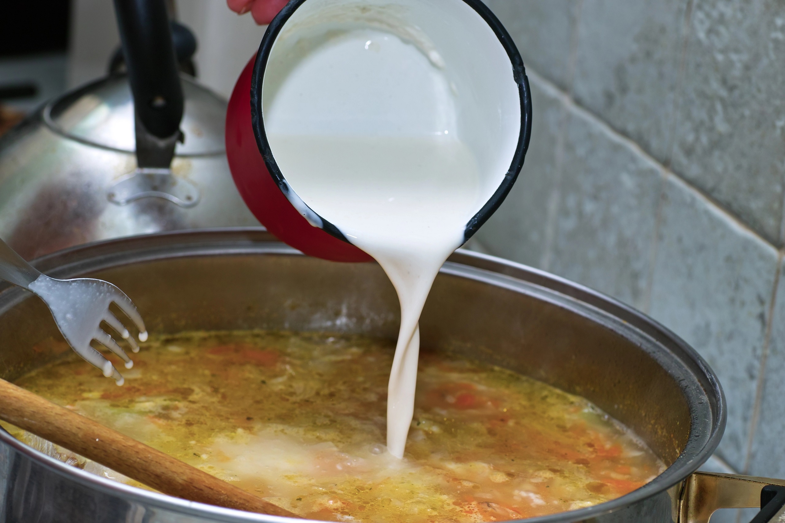 Milk or cream being poured from a red saucepan into a simmering steel pot of pickled cucumber soup to thicken it at the perfect moment with a big wooden spoon for stirring.