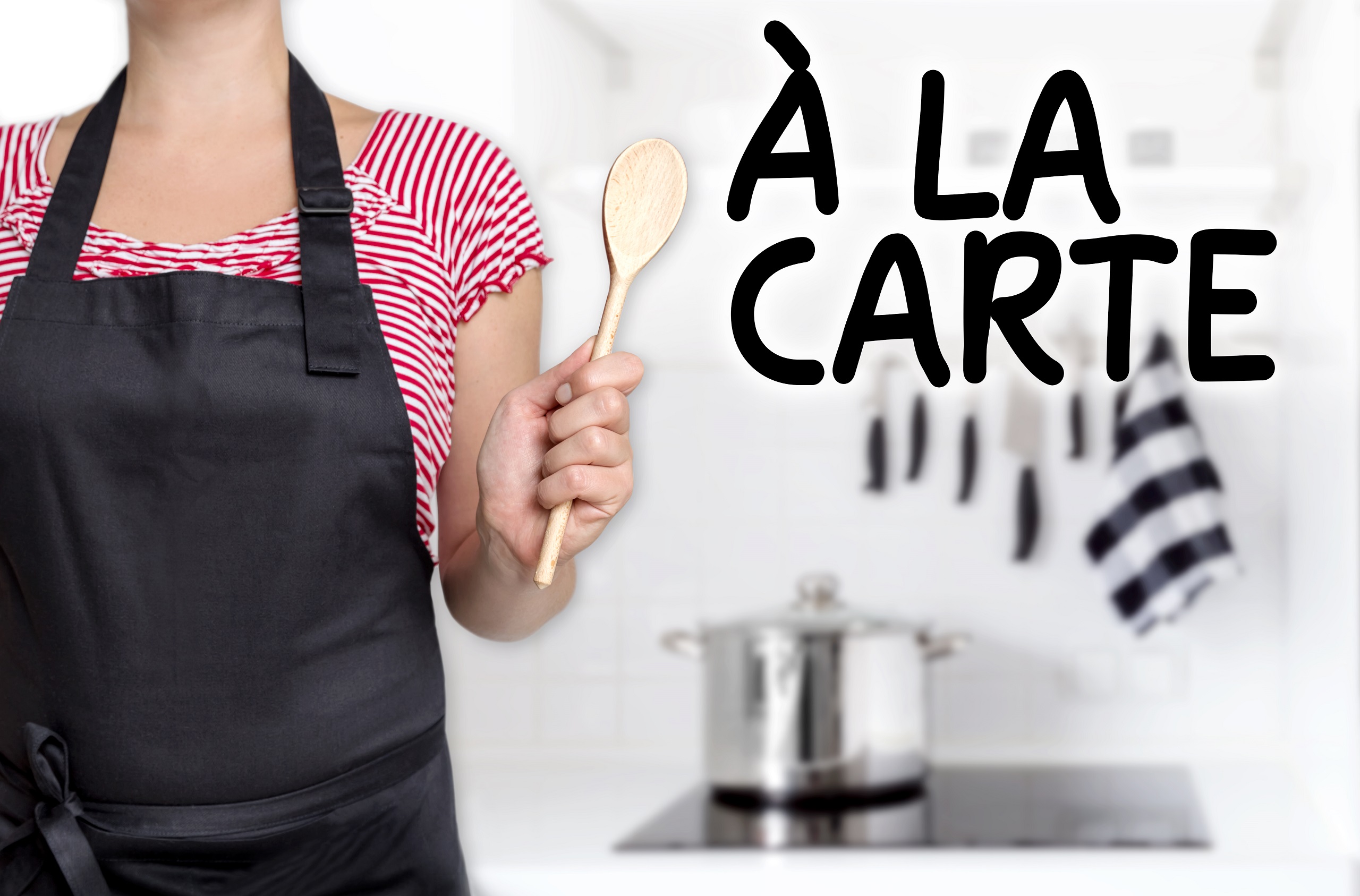 A female in a red and white short sleeved shirt wearing a black apron holding a wooden spoon angled towards the words "a la carte" with a kitchen background in a white setting.