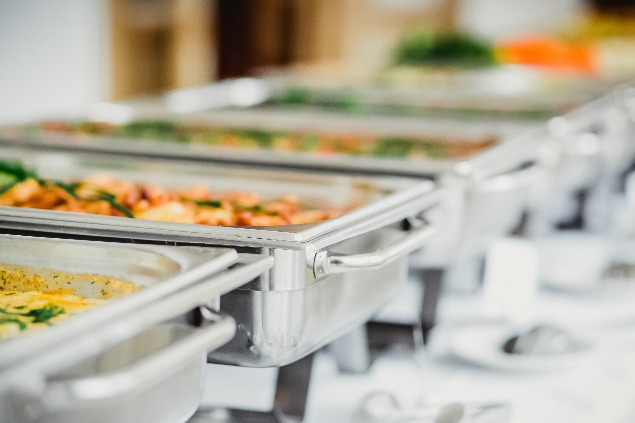 Buffet food trays with food neatly arranged in a row on a long table adourned in an elegant white cloth and several plates with silverware depicting a catered food event such as a wedding or birthday.