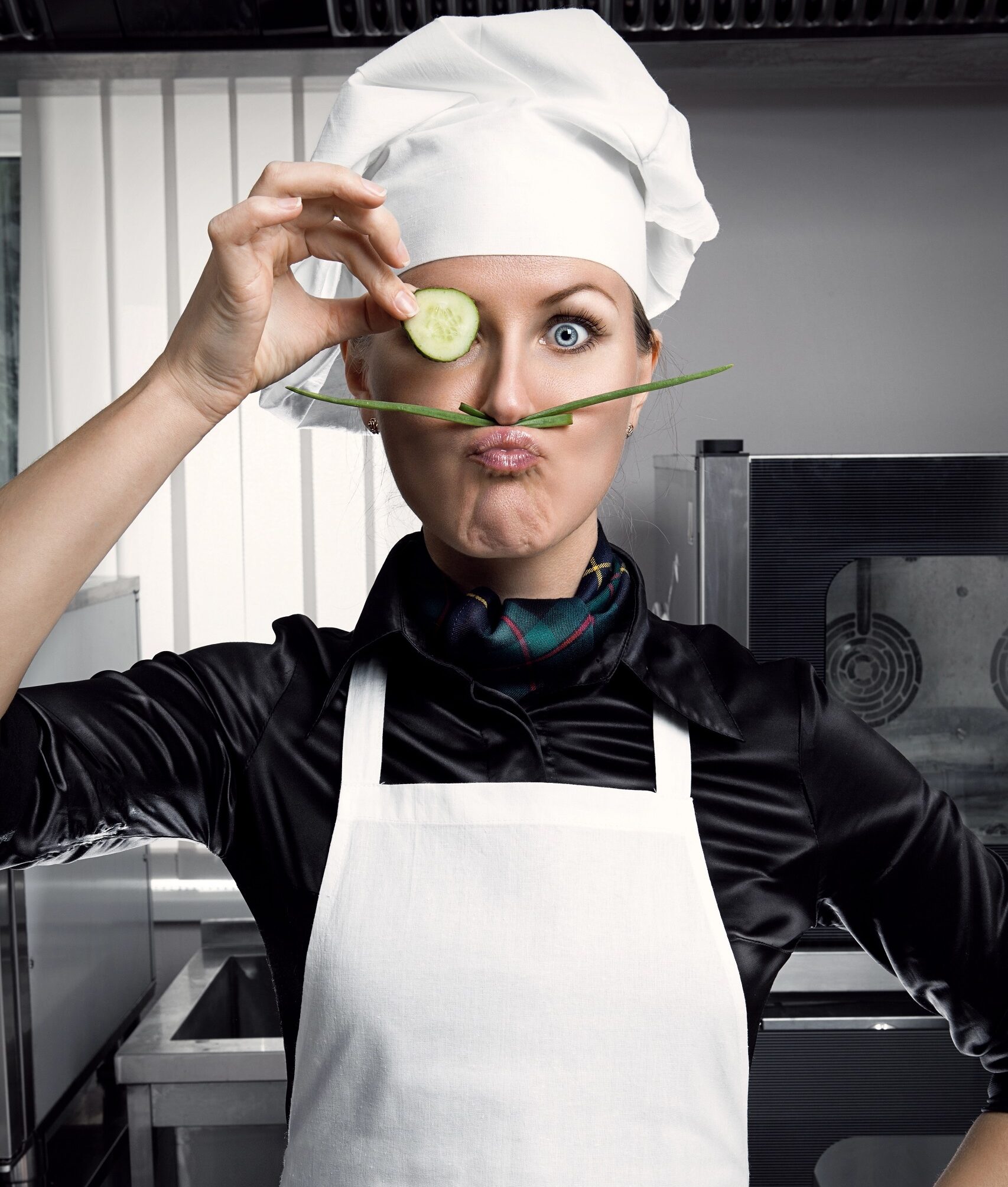 Pretty female wearing a white chef's apron and hat with a black shirt underneath posing with one hand on the hip and the other holding a sliced cucumber to her eye with her forefingers and an asparagus held up between her upper lip and nose as if a moustache, depicting her silly good nature, with a commercial kitchen in the background.
