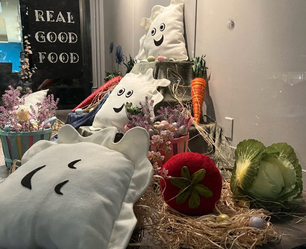 Kat's Restaurant window display with several life-sized handmade pierogi pillows making different facial expressions, large handmade vegetable pillows such as carrots, cabbage, and beets, all laid out on a bed of real straw in an inviting arrangement with a handmade sign hanging in the background made from white caligraphy lettering on a black canvas having the words real good food.