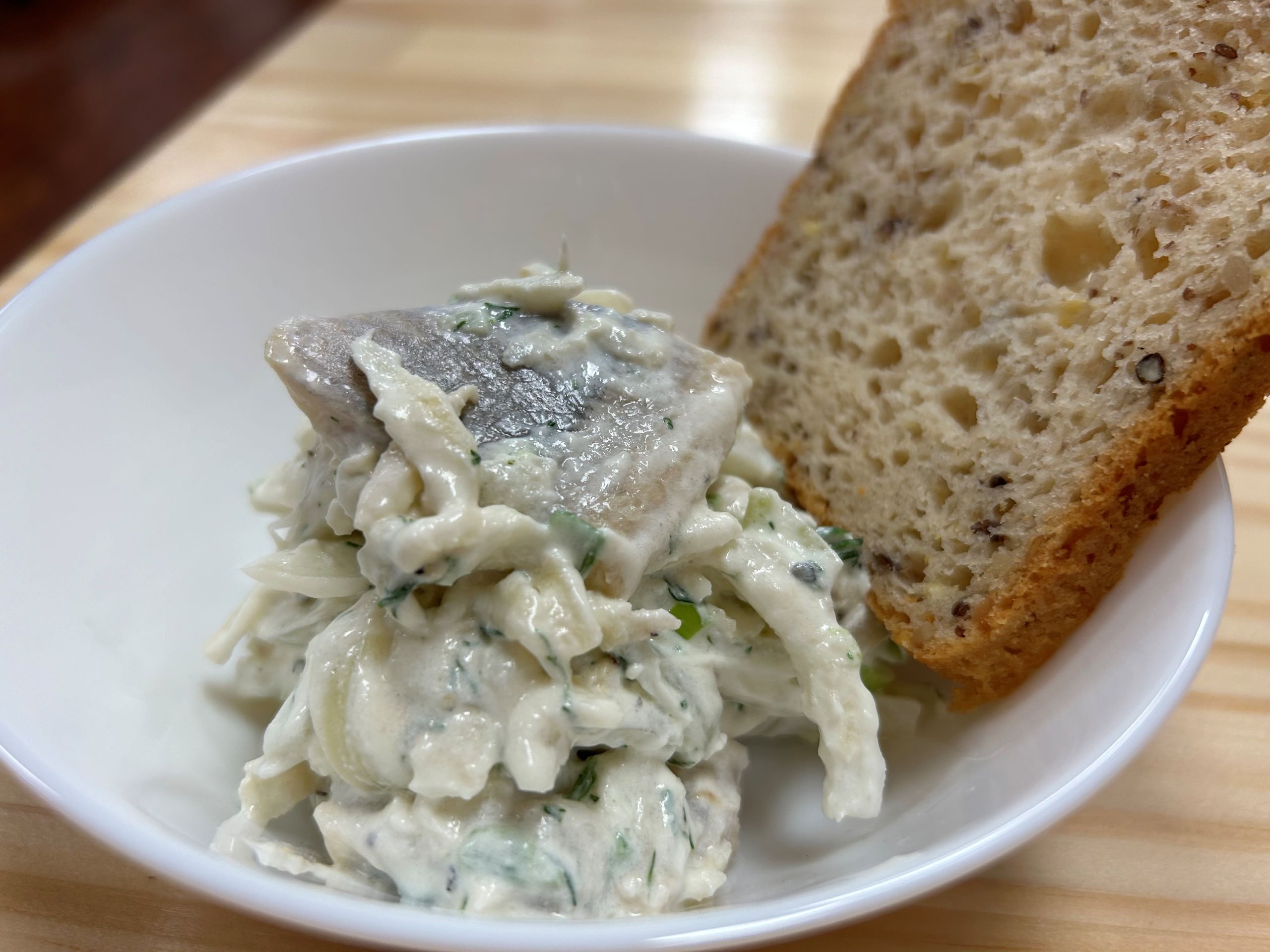 Kat's Creamy Herrings with Dill Sauce having pieces of marinated herring fillets and white creamy sauce stacked in a white dish with a slice of multi grain bread on an ash wood table.