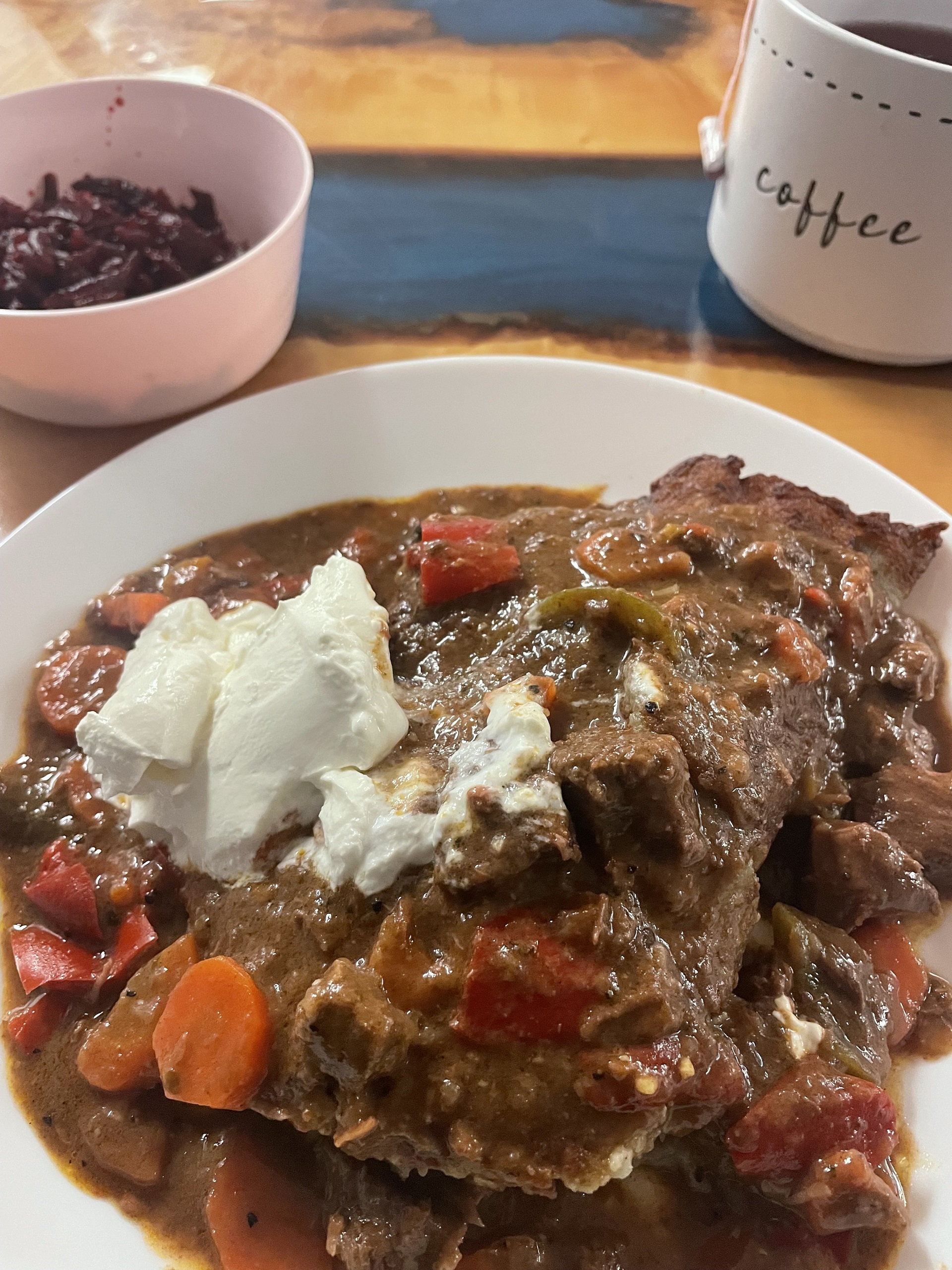 Kat's Goulash Beef Bleenie with tender savory beef chunks and vegetables such as sliced carrots, pieces of red paprika, served in and on an XL bleenie folded with beef goulash gravy and a dollop of sour cream on a white plate with a side of Slavic veggie beet or red cabbage salad on white dishes.