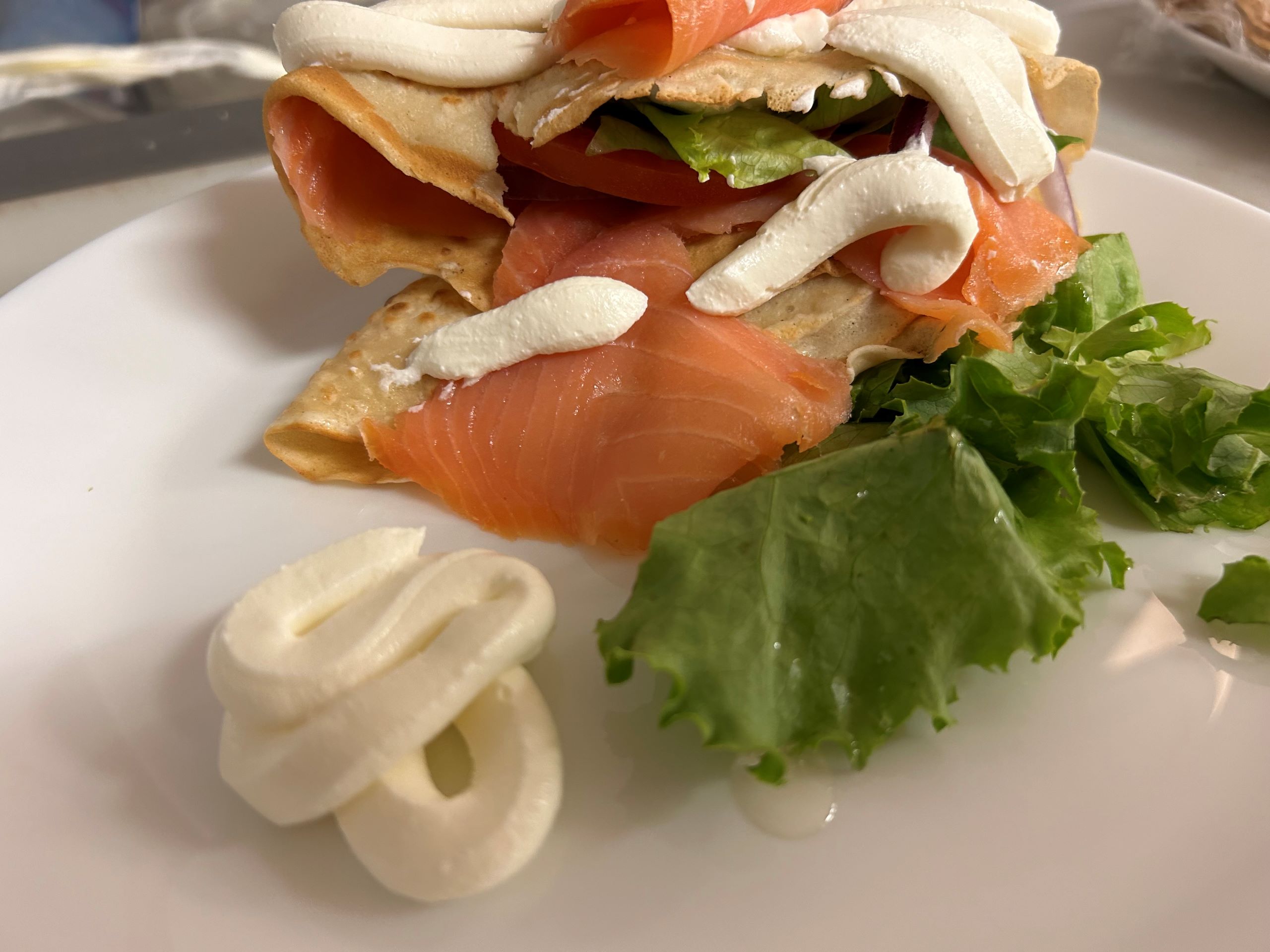 Slices of Smoked Salmon and Cream Cheese Spread artfully wrapped in a blintz pancake with crispy lettuce and slices of tomatoes on a white plate.