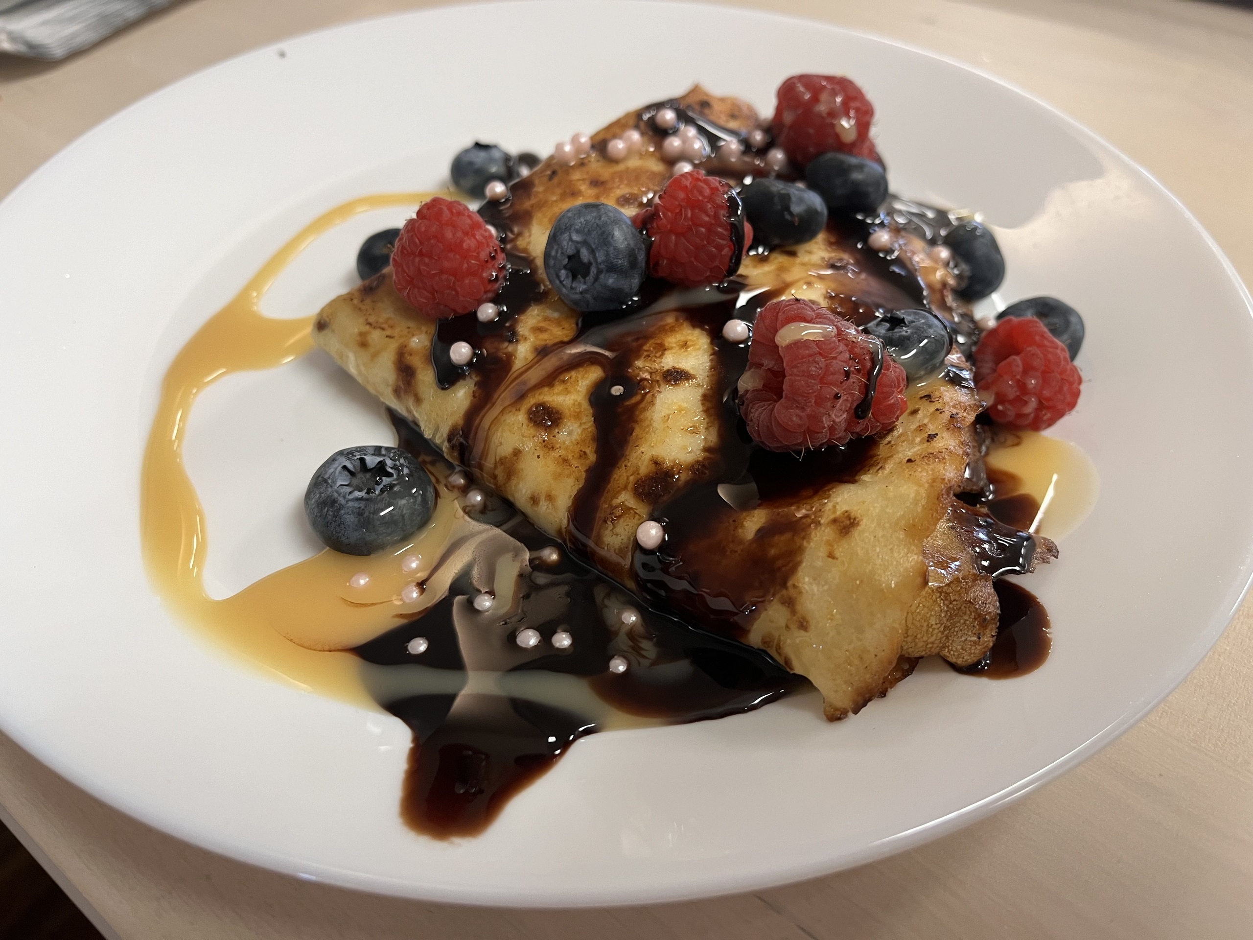 Kat's sweet cheese blintzes topped with blueberries, rasberries, and edible sugar pearls, and homemade chocolate sauce on a white plate on an ash wood table.