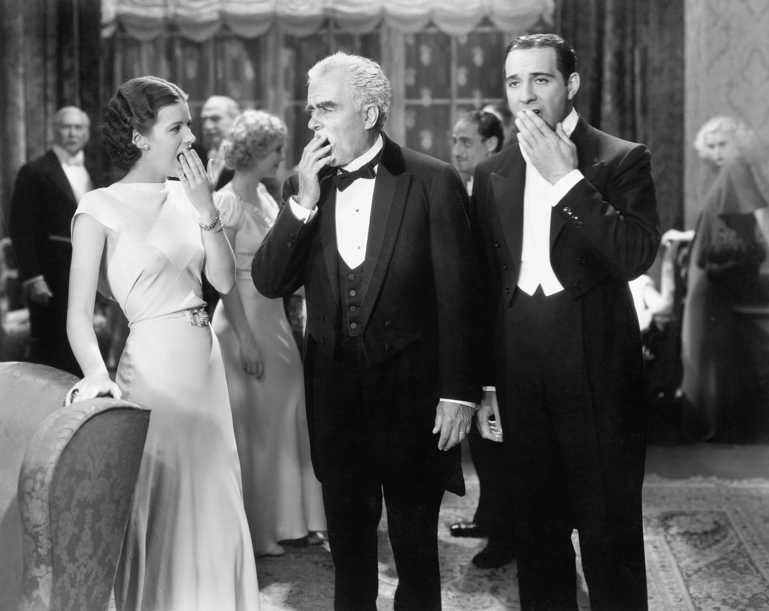 Two men in tuxedos and a lady in a gown standing and yawning at the forefront of vintage white and black photo with other people wearing formal wear mingling in the background depicting an early 20th century elegant party.