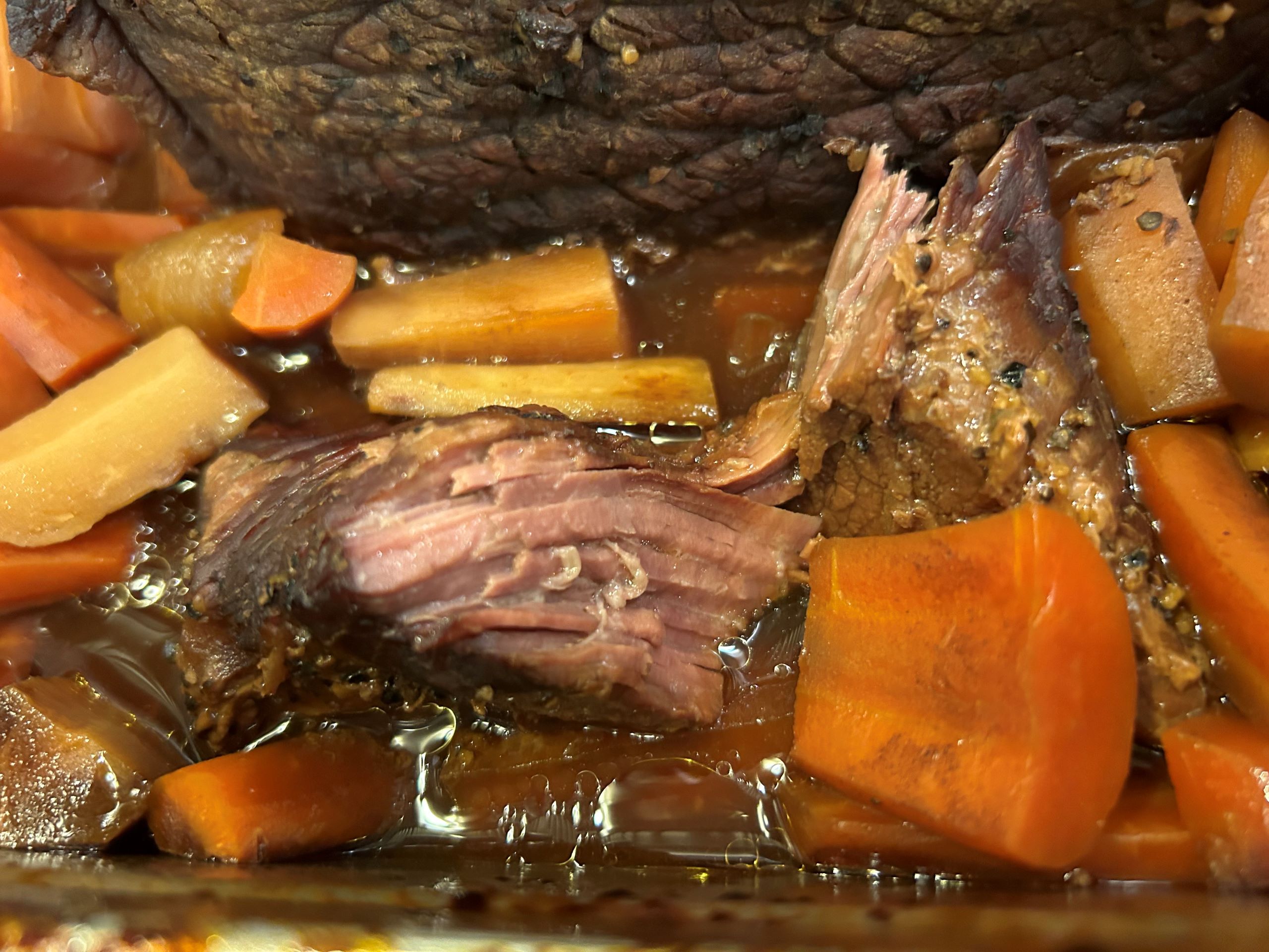 Kat's Baked Brisket right out of the oven with the juices still bubbling and vegetables pieces such as cooked carrots, garlic, onions, peppercorn seasoning, and a chunk of tender meat showing the stringy goodness and right color of the brisket meat when cooked.