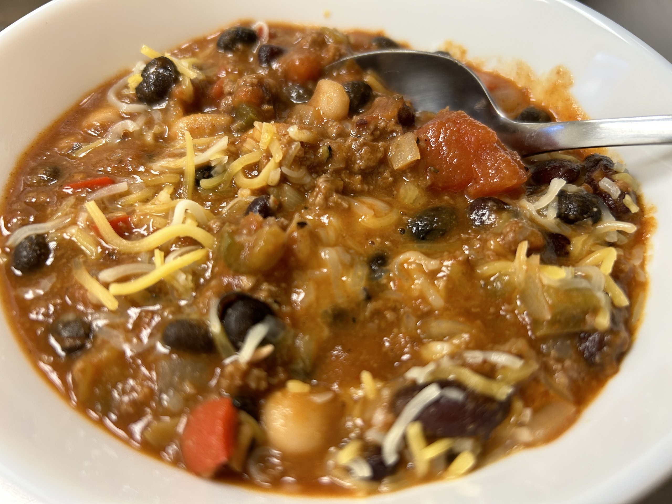 A close up of Kat's chilli bean soup in a white bowl with black beans, ground meat, chunky stew tomatoes, shredded cheese, in natural tomato and chilli juices with a spoon resting in the bowl.