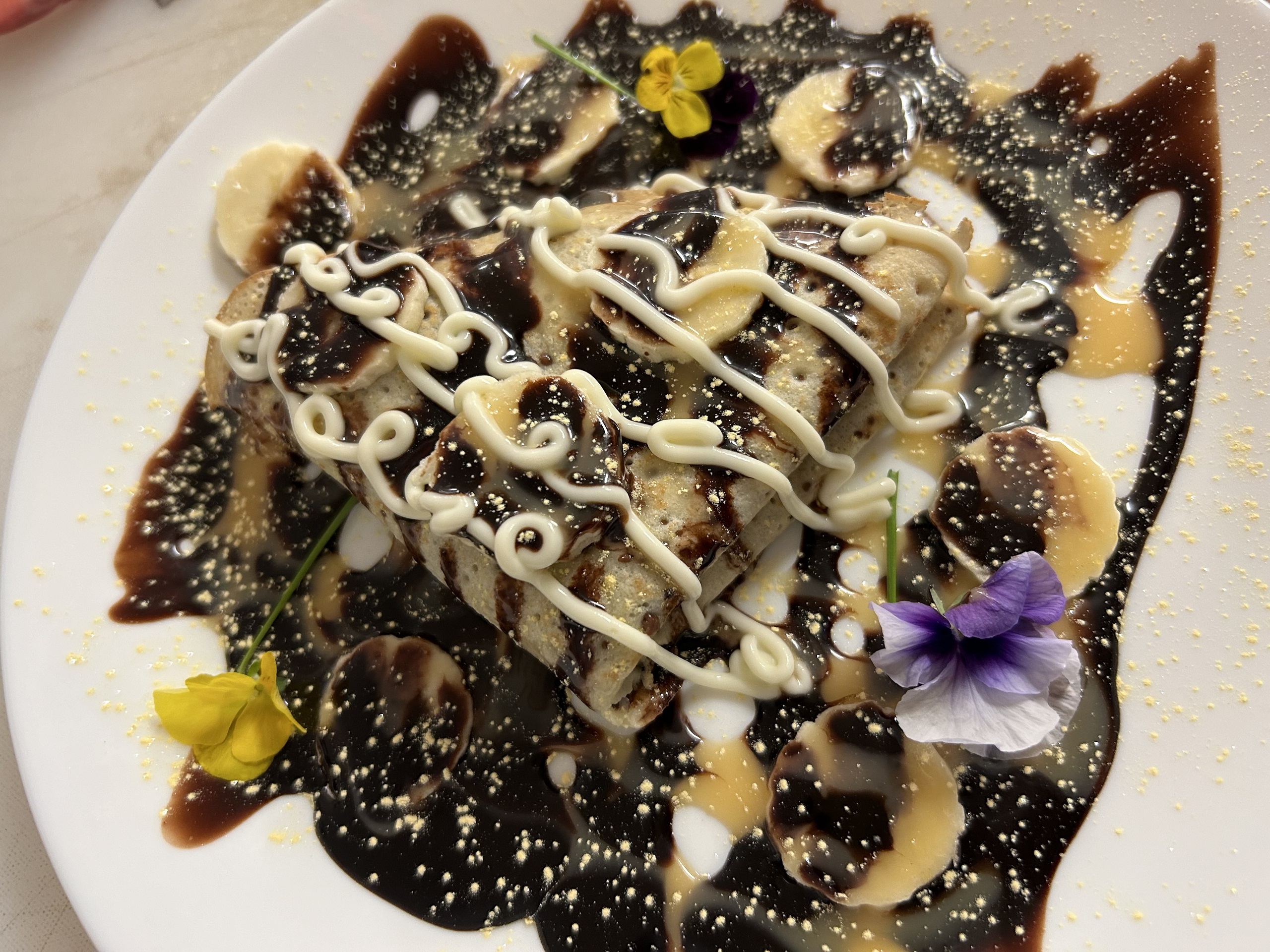 Kat's Nutella and Banana Blintz covered in chocolate sauce and sliced bananas with a swiggle of sweet cream on a white plain garinshed with edible flowers.