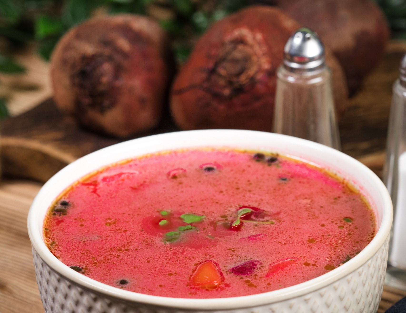 Red beet borscht soup with potatoes and herbs in a white bowl thickened with sour cream and having whole beets and green with salt and peper shakers in the background.