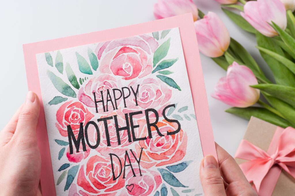 Happy Mother's Day Card having bold black letters with the words "happy mother's day" in the middle of a white card with pink tulips and green leaves artwork being held by a woman's hands on top of a pink envelope with real tulip flowers resting on a white background with a small present in a brown box wrapped in a pink bowtie ribbon.