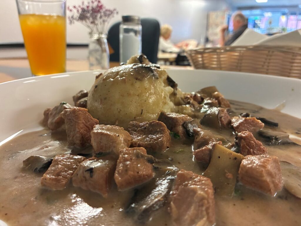 Kat's Beef in Mushroom Sauce and Mashed Potatoes on a white plate with the restaurant in the background including a pair of customers and items on the table such as an orange-mango juice in a glass, saltshaker, flowers, napkins, and a small bamboo basket.
