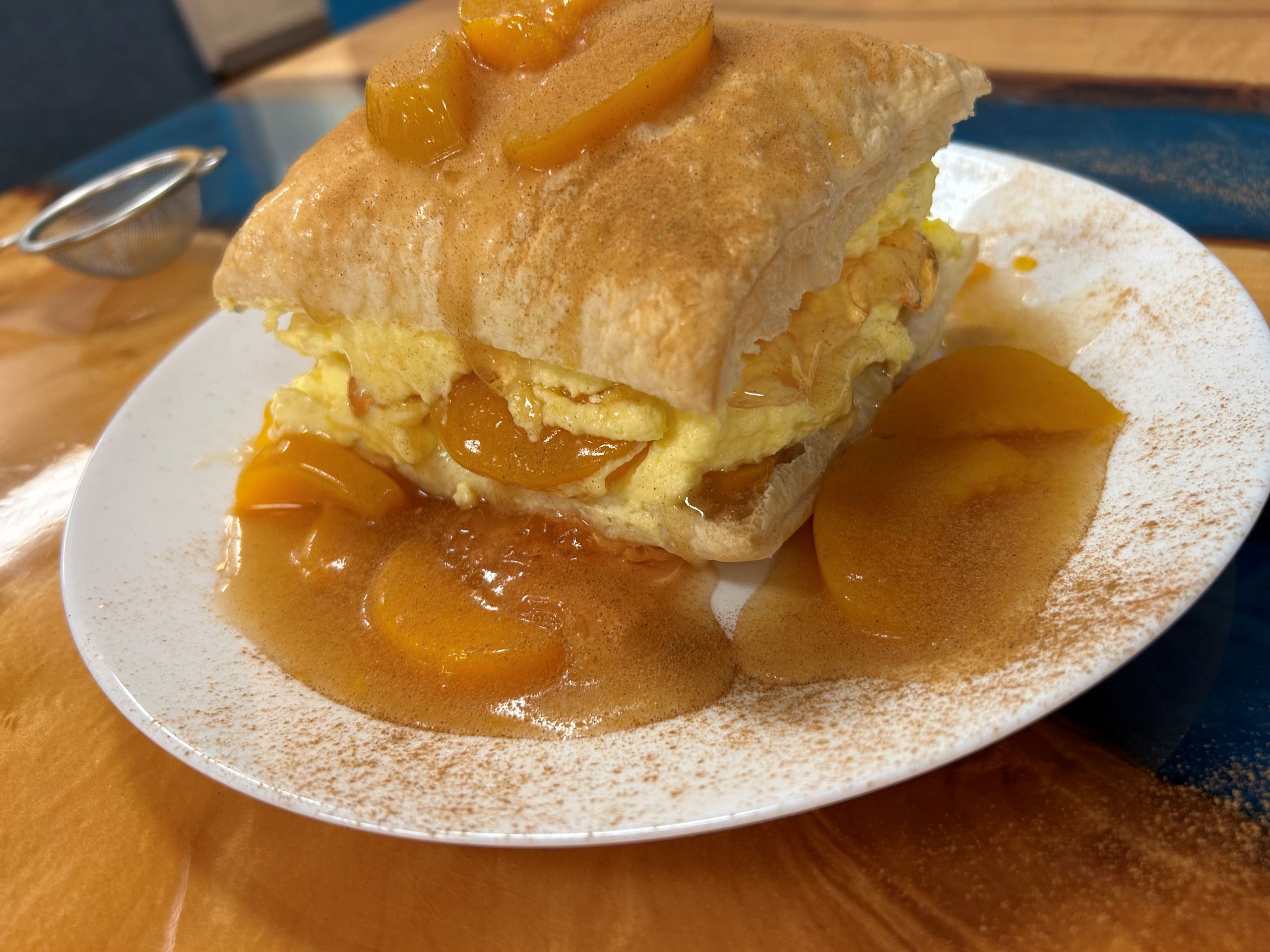 Kat's Slavic Napoleon dessert made from flakey french pastry croissant-like dough filled with light cream, peaches, and peach jello, topped with more peaches and sprinkled with cinnamon served on a white plate with peach syrup, peaches and garnished with cinnamon, on the custom ambrosia maple wood table at Kat's Cafe in Minersville.
