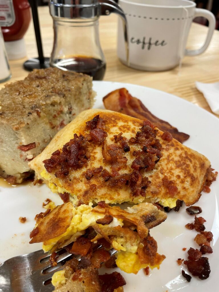 Kat's Bacon Egg and Cheese Blintz partially eating with bacon bits and bacon strip there being a metal fork with a mouthful on it and Potato Babka with maple syrup on a white plate on an ash table with a white mug with the word "coffee" and a maple syrup glass pot.