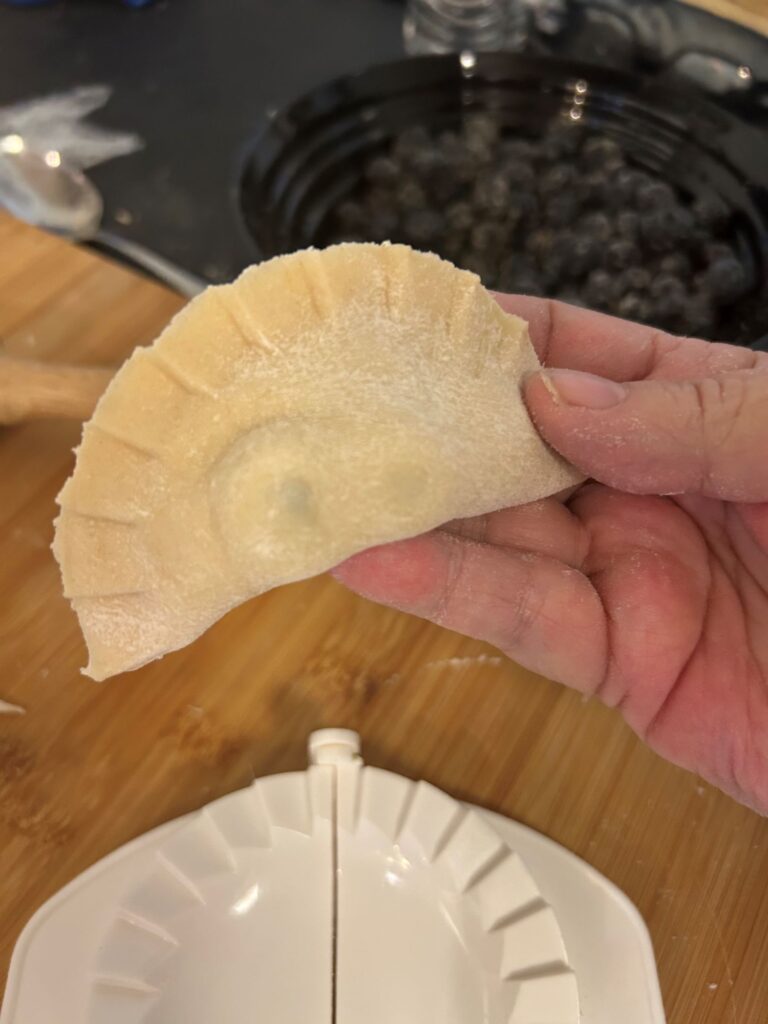 Kat's Hand Crimped Large Pierogi Made from Scratch Being Held up by a Female Hand and below it you see the single pierogi hand crimp utensil and a bowl of blueberries, as well as a wood grain butcher block and a stainless steel surface.