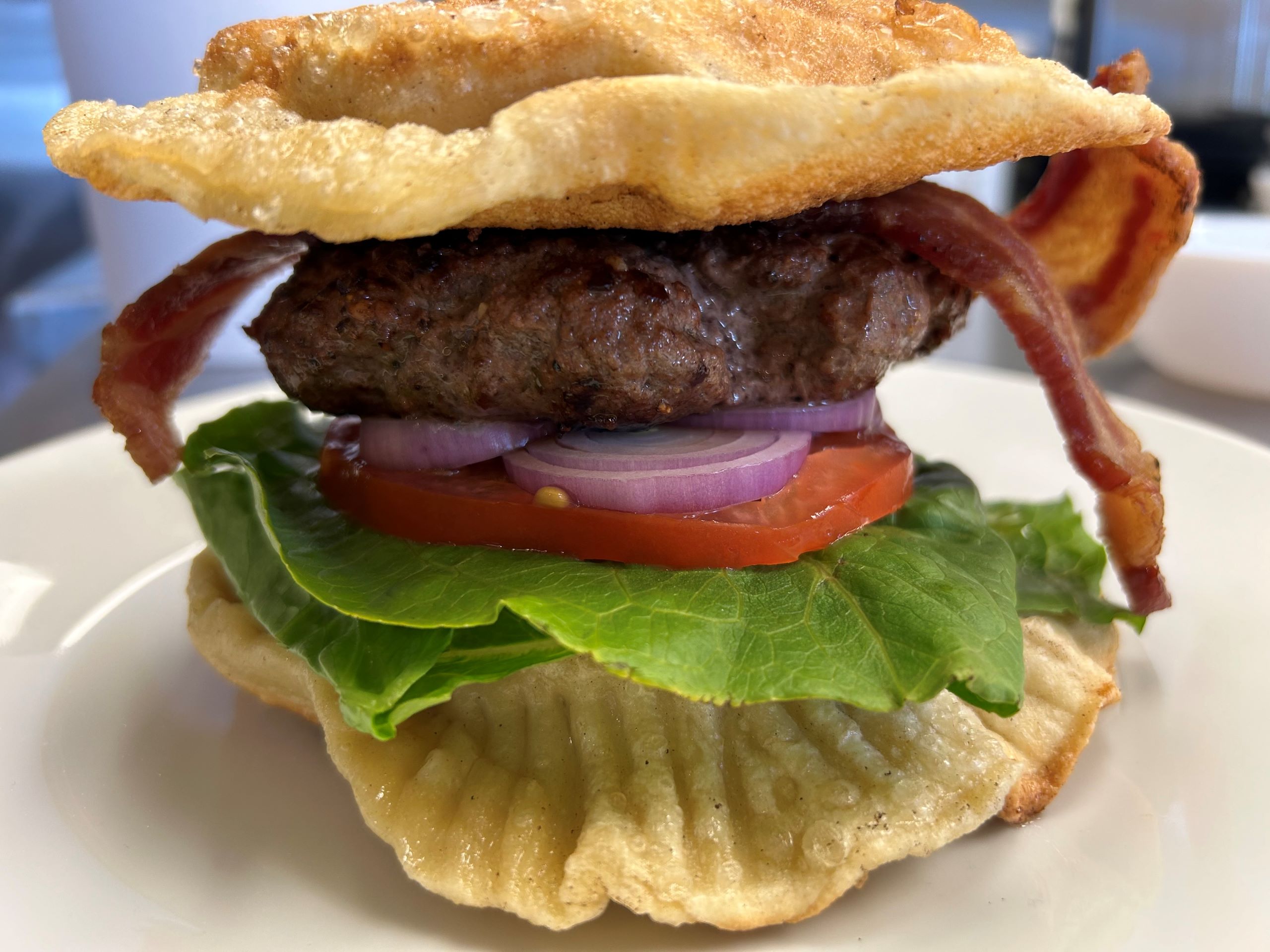 Kat's Pierogi Smash Burger is 2 XL hand-made crispy potato-cheese pierogies sandwiching a smash patty burger made from scratch with strips of bacon, sliced onions, sliced tomatoes, and lettuce, served on a white plate.