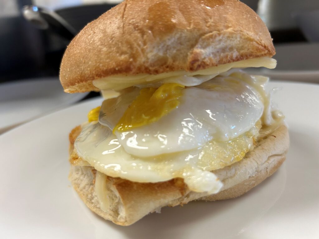 Kat's classic breakfast sandwich having a couple of fried eggs and a slice of cheese on a fresh, lightly toasted, breakfast roll, served on a white plate.