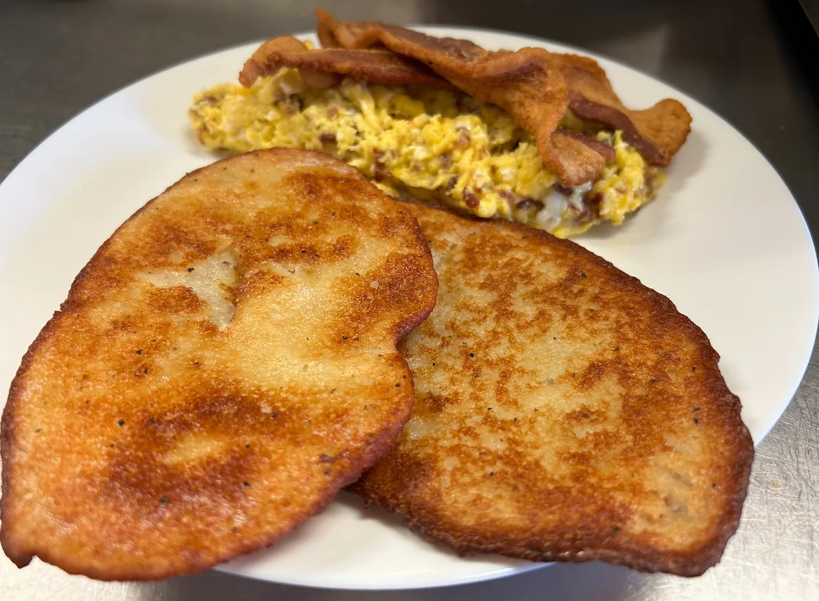 Two potato bleenies with browned, crisp edges in the foreground, with scrambled eggs and bacon behind them on a white plate at Kat’s.