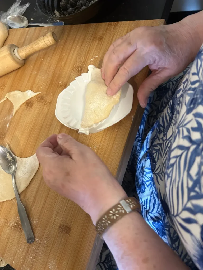 Hands folding and sealing a blueberry pierogi in a single hand press on a floured wooden work surface in the kitchen Kat’s Café & Restaurant.