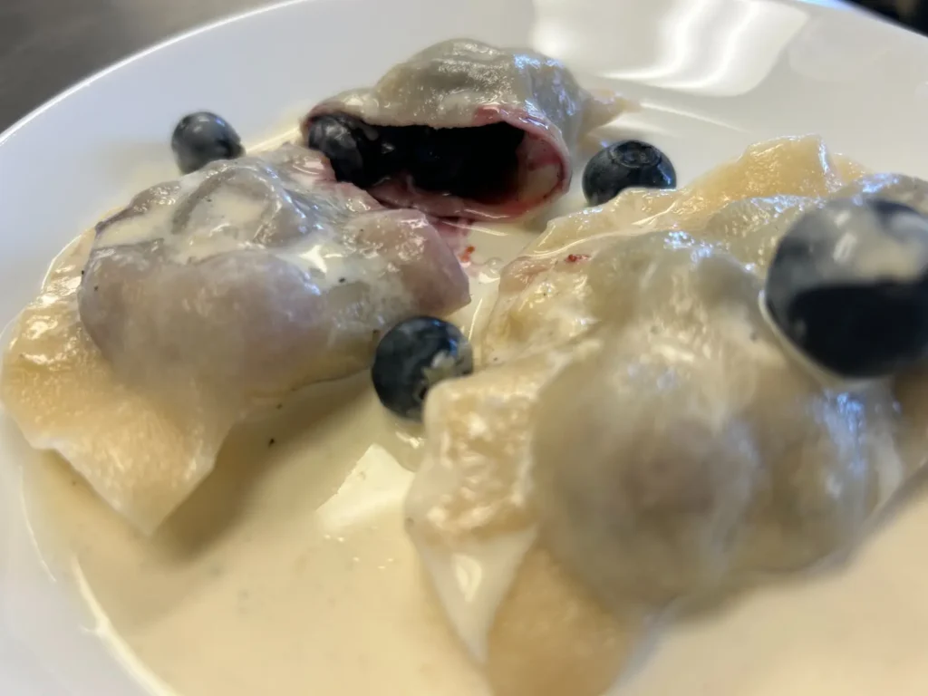 Close-up of handmade blueberry pierogies in light sweet cream, including one opened to reveal its fresh blueberry filling, with whole blueberries on the plate at Kat’s Café & Restaurant.