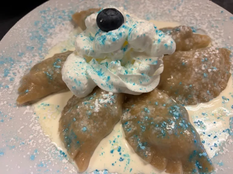 Close-up of handmade blueberry pierogies served with cream, whipped cream, powdered sugar, blue decorative sugar, and a fresh blueberry at Kat’s Café & Restaurant.
