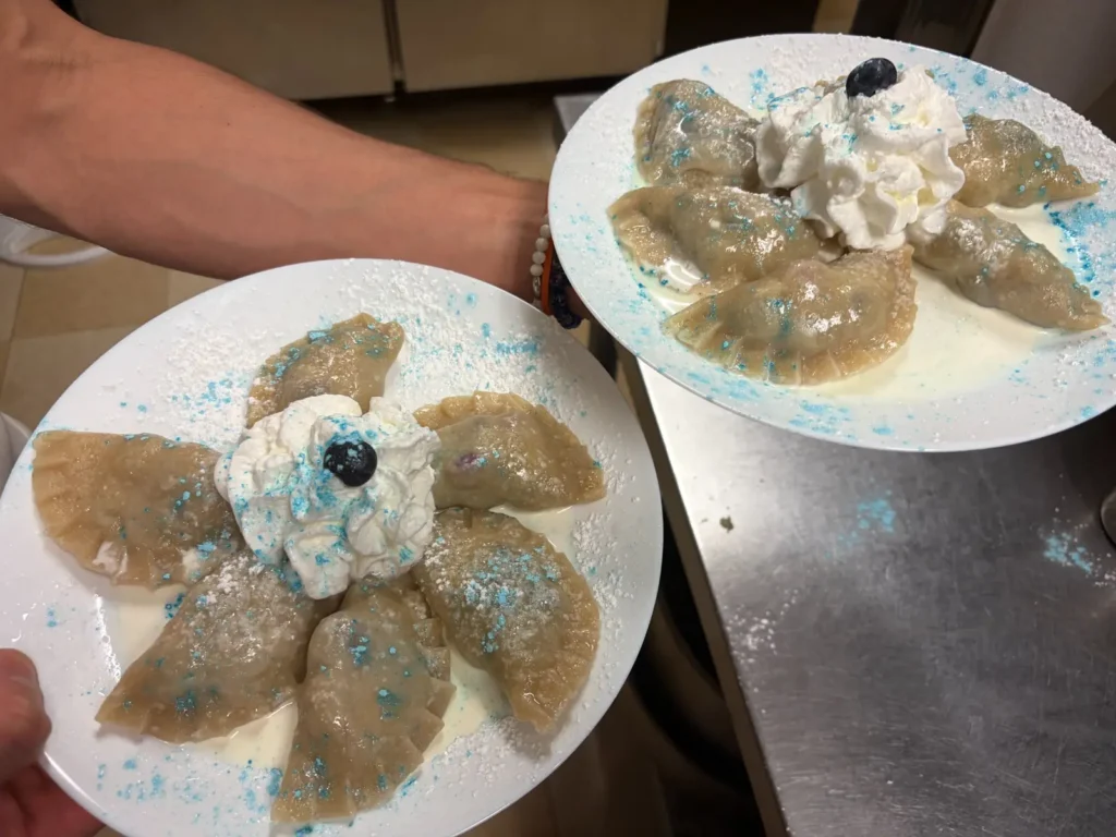 Two freshly prepared plates of handmade blueberry pierogies with cream, whipped cream, powdered sugar, blue decorative sugar, and blueberries held in the kitchen at Kat’s Café & Restaurant.