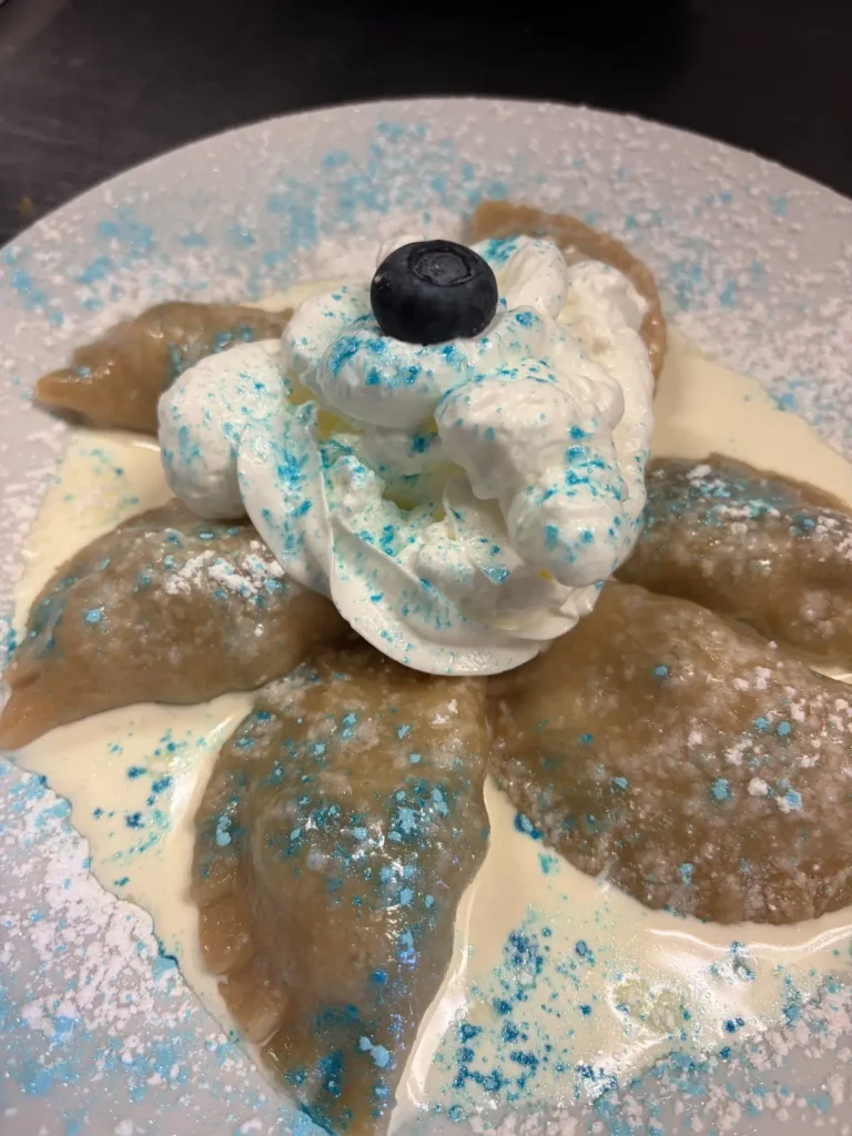 Close-up of seasonal handmade blueberry pierogies served with cream, whipped cream, powdered sugar, blue decorative sugar, and a fresh blueberry at Kat’s Café & Restaurant.