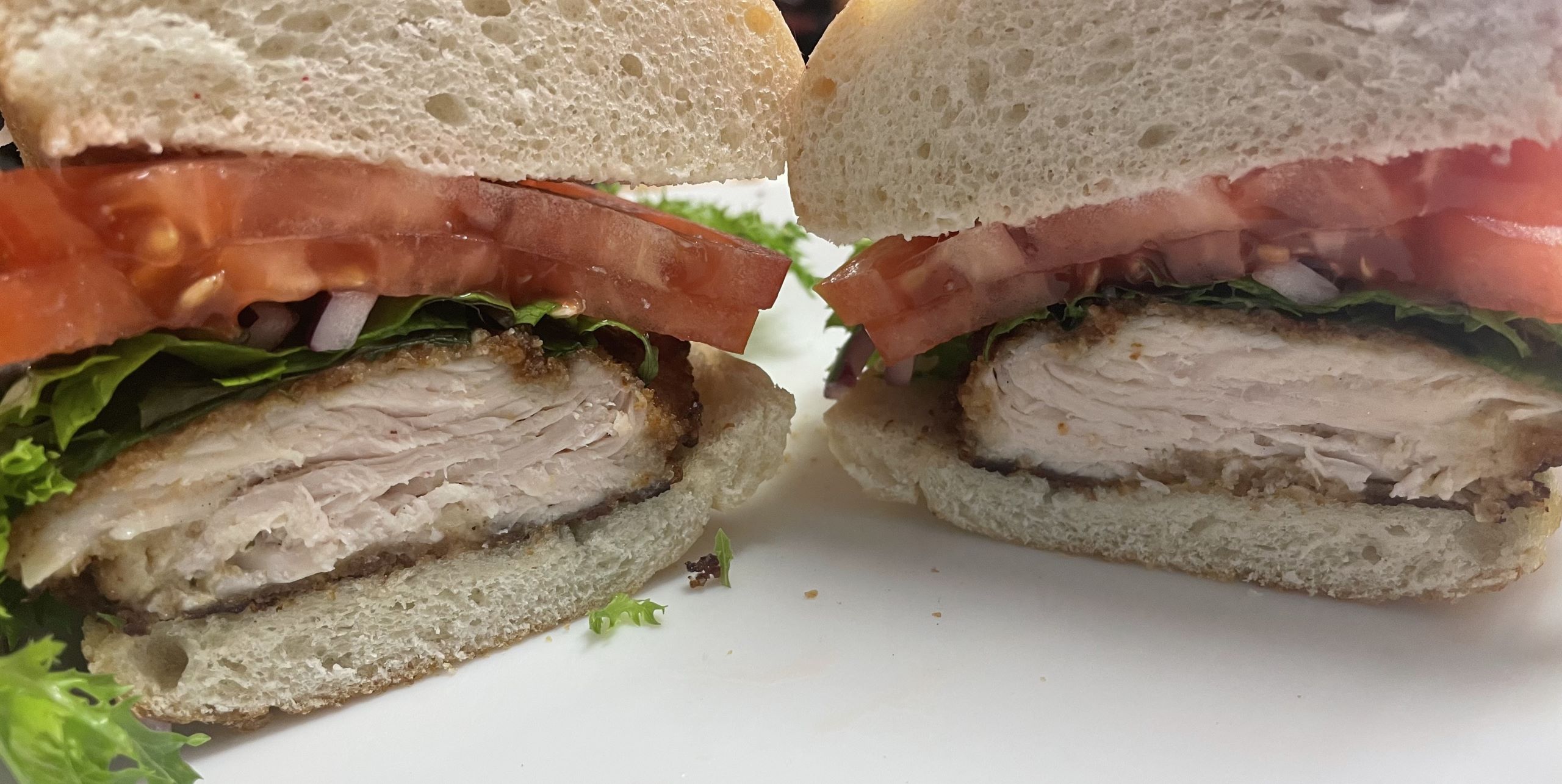 Kat's Chicken Cutlet Hoagie cut in half showing both halves having tender natural free-range chicken breaded with generous slices of tomato, lettuce and red onion on white submarine bread, served on a white plate.