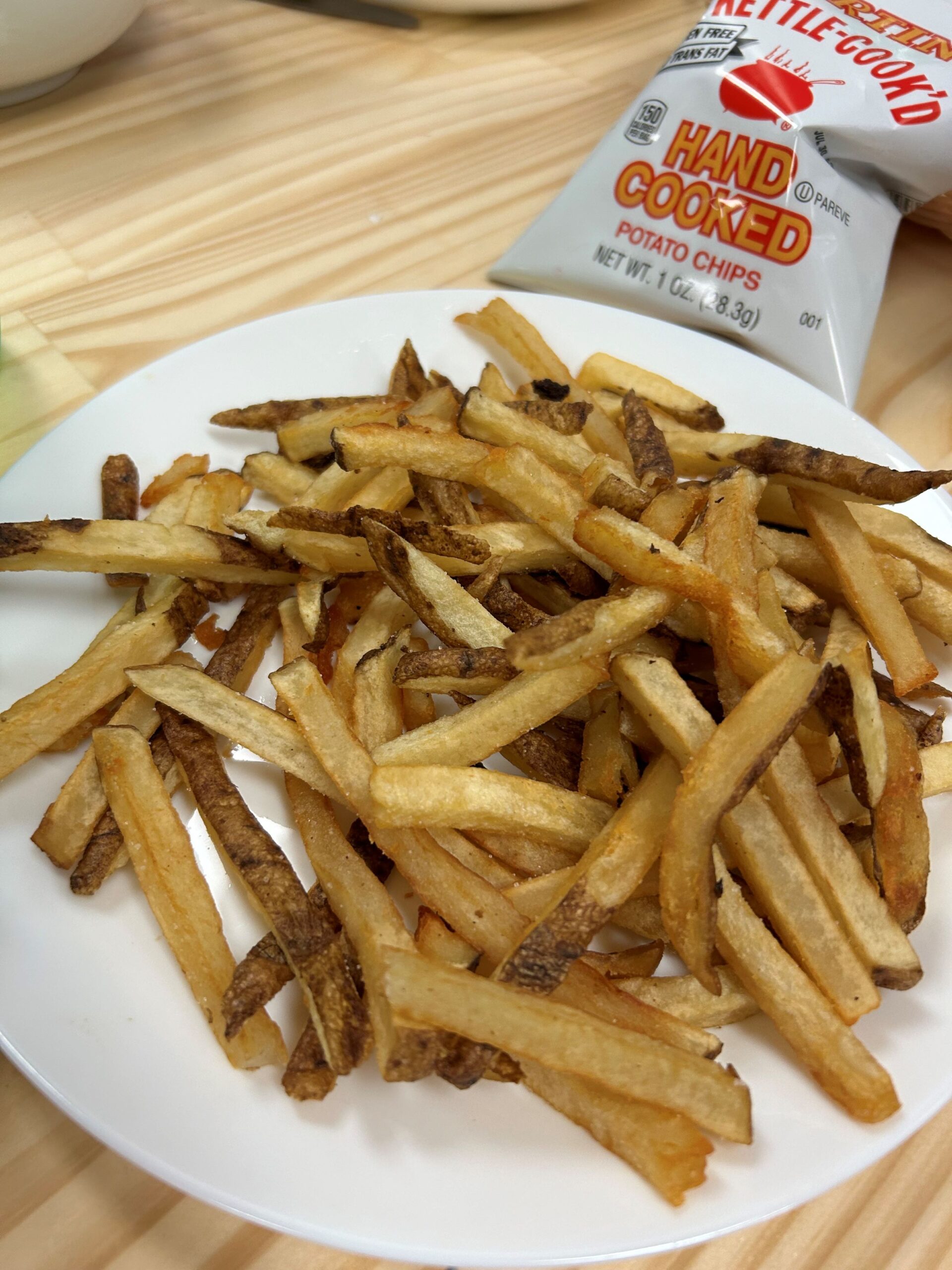 Kat's Fresh Cut Fries from real Idaho potatoes served on a white plate having a bag of potato chips placed next to it on an ash wood table.