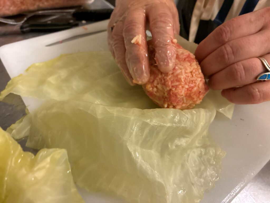 Kat's Fresh Halupki Filling Being Rolled into a XL Cabbage Leaf by a Female Pair of Hands on a White Cutting Board on a Stainless Steel Surface with a Bin of Meat Filling and Kitchen Caterware.