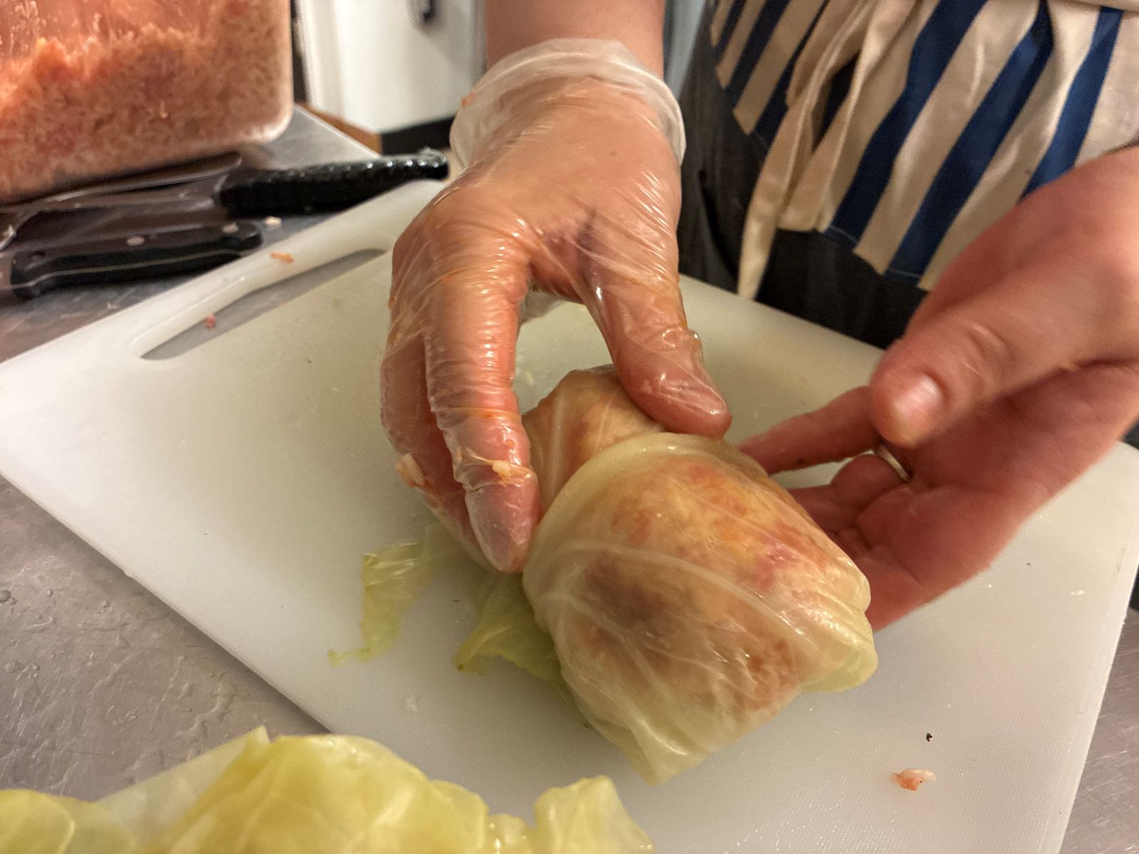 Kat's Fresh Halupki Gołabki Just Rolled into a Cabbage Leaf and Being Lightly Held Up by a Pair of Female Hands with the Bottom Portion of the Halupki Gołabki Resting Against a White Cutting Board Underneath it on a Stainless Steel Surface with a portion of the cabbage leaf and a bin of meat-rice filling also adjacent the kitchen work station.