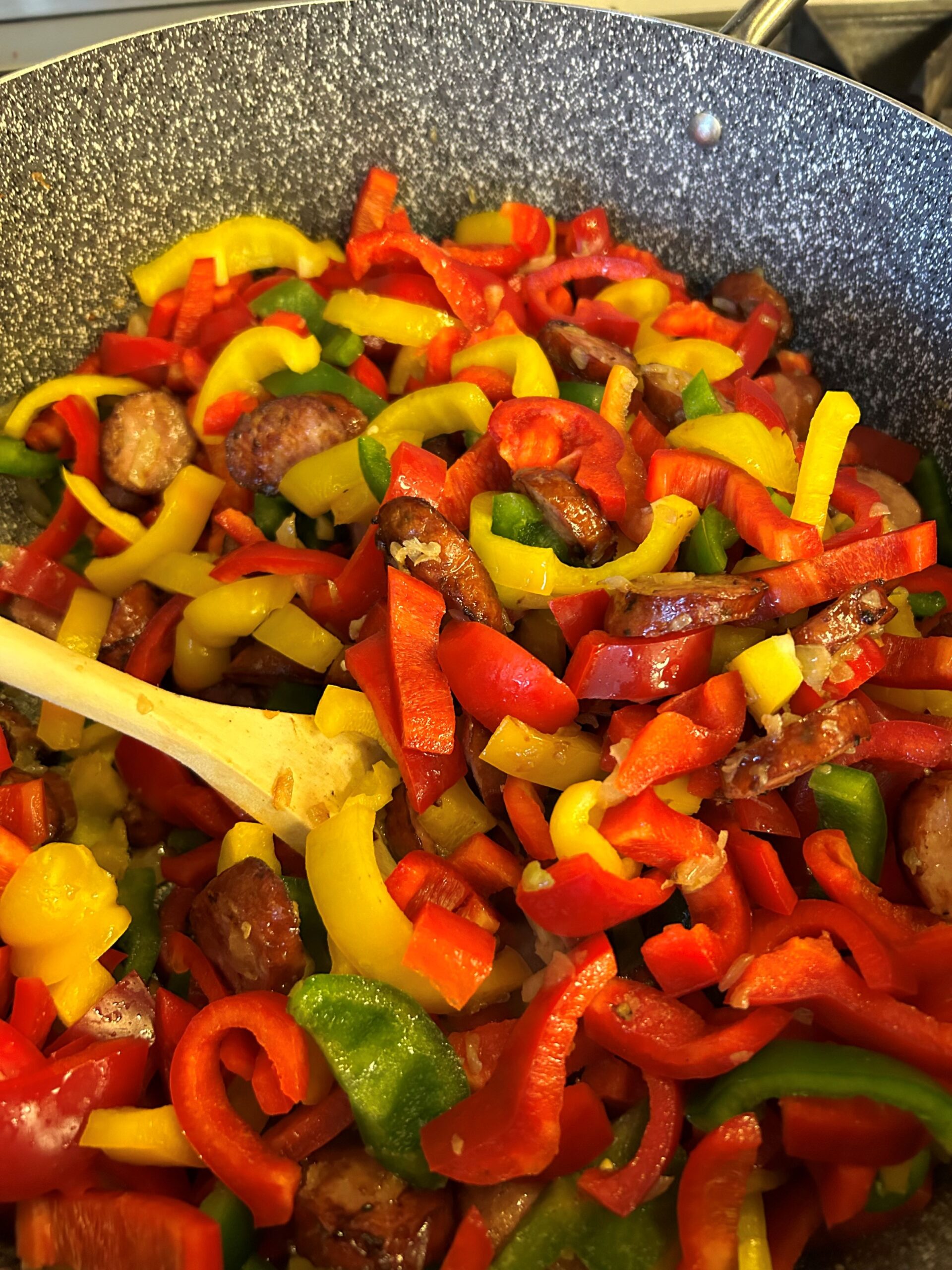 Kat's Letcho Kielbasa Fresh Peppers Being Made in a saute pan mixed with kielbasa pieces and onions being stirred with a wooden spoon.