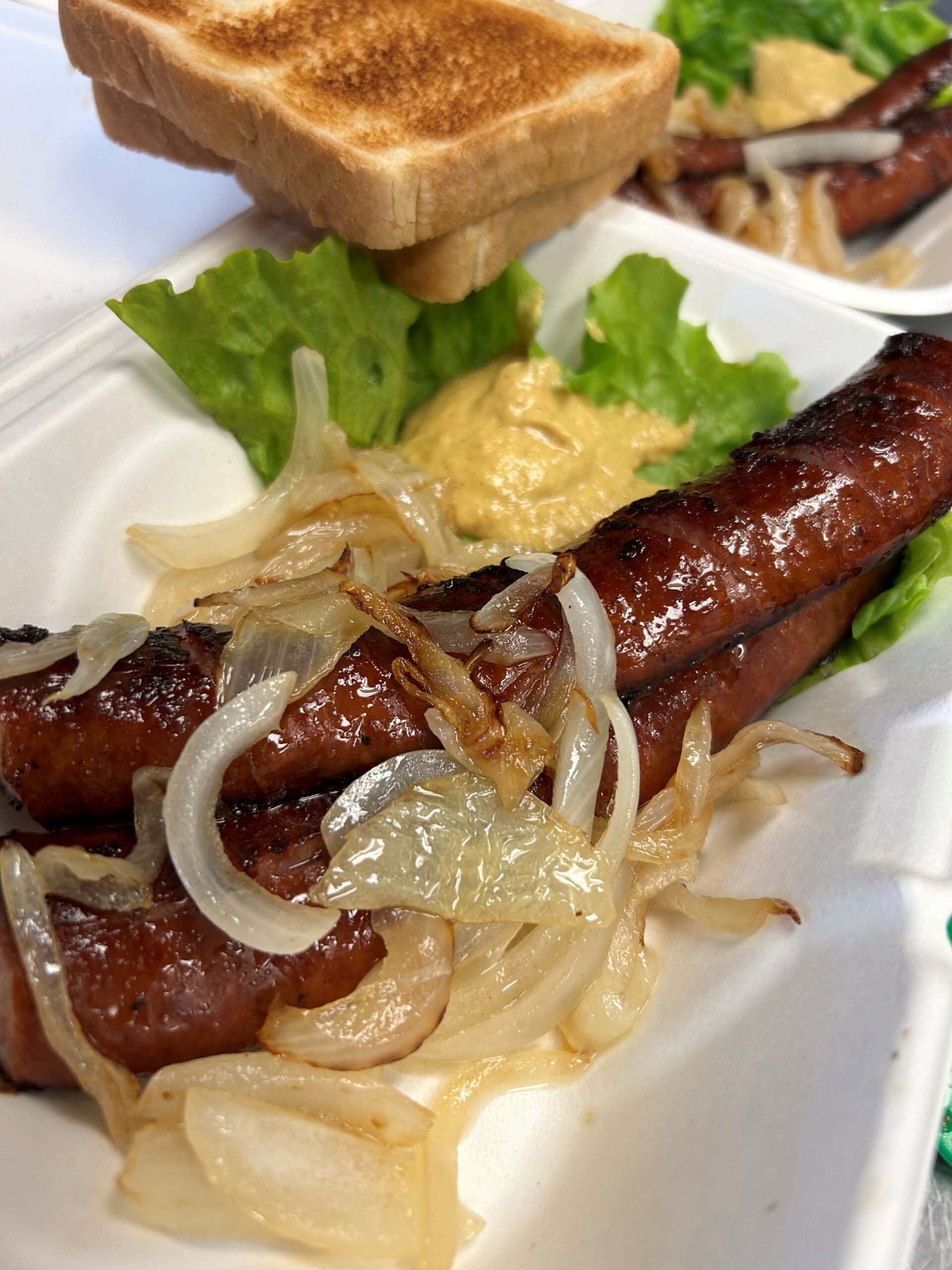 Kat's Polish Kielbasa Appetizer Takeout Order with long slivers of kielbasa, topped with fried onions, having a serving of Slavic mustard on a bed of greens, and slices of toast in a white take-out food container with another kielbasa order in the background.
